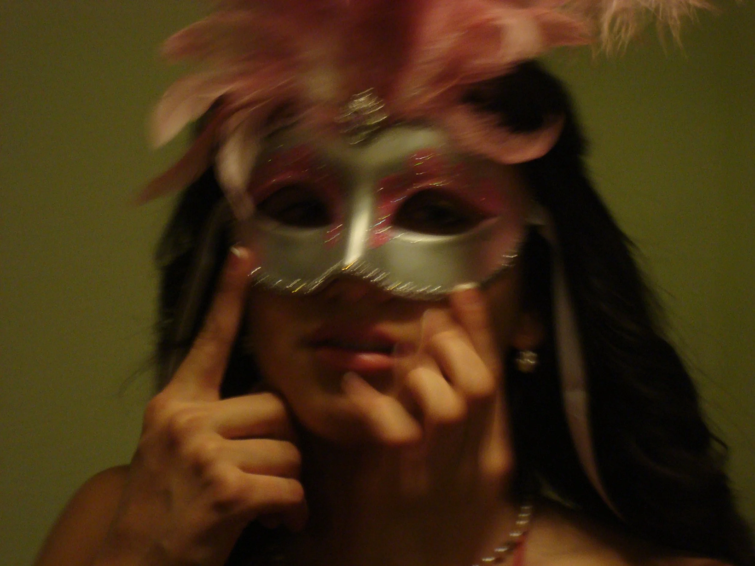 A person wearing a silver mask with red and black accents, holding the mask with their fingers, with long dark hair and a pearl necklace, against a green background.