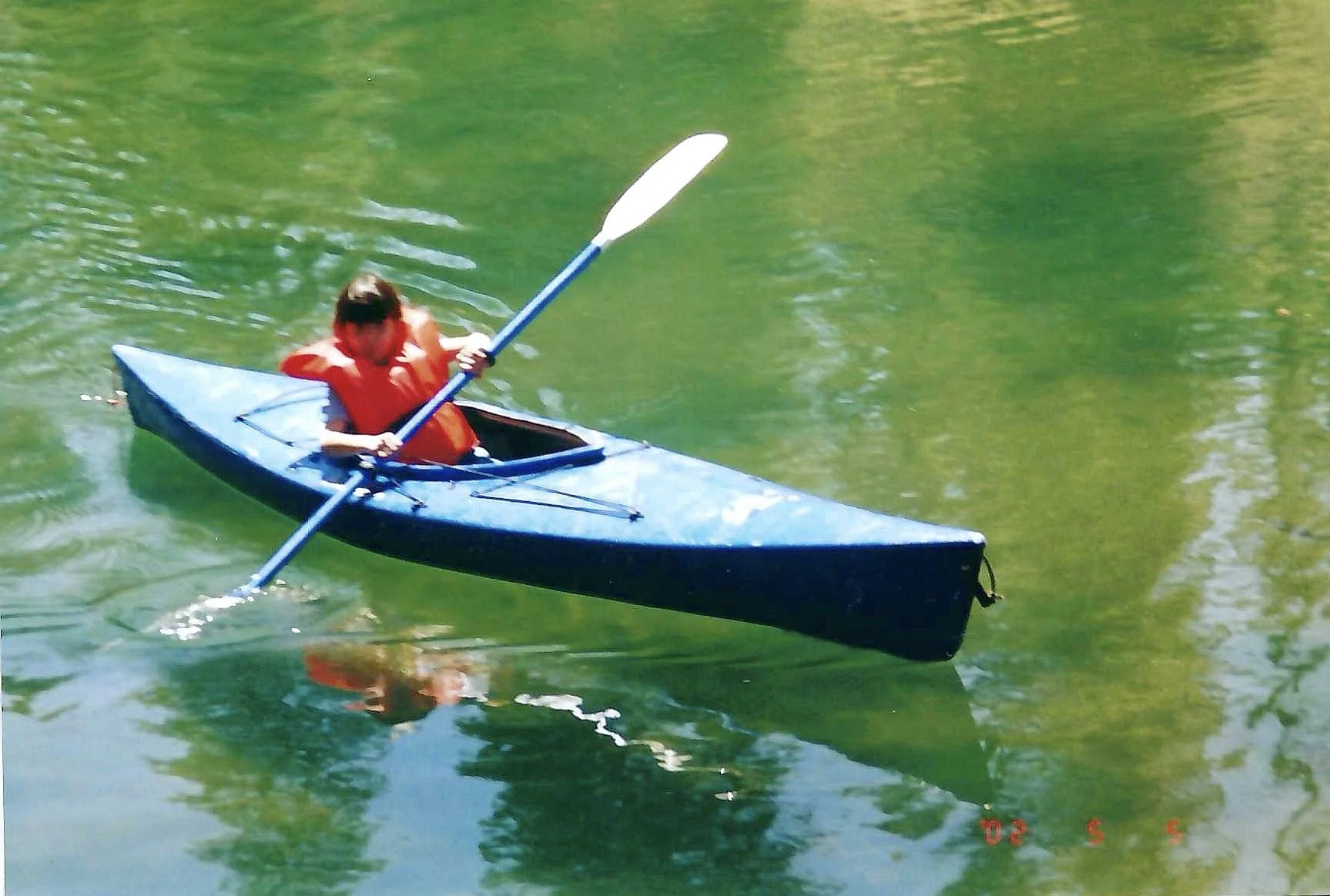A person in a red life jacket paddling a blue kayak on a green lake.
