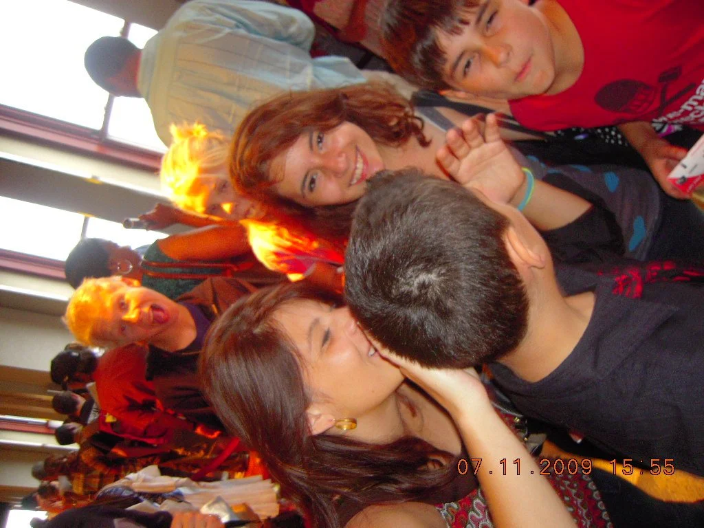Group of people at a social gathering, with two women and one man engaged in a close conversation in the foreground, smiling and making eye contact, while other people are in the background in a room with large windows.