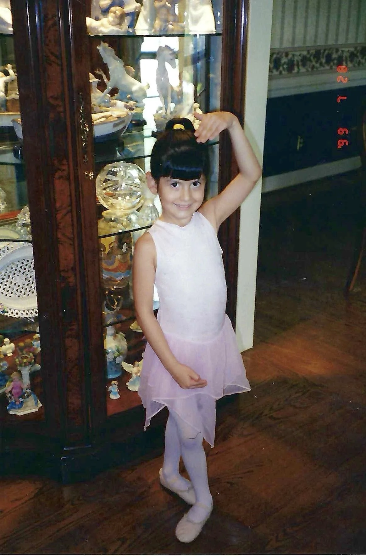 A young girl with black hair and a high ponytail in a yellow hair band, wearing a pink dress and white tights, posing with her right arm curved over her head and her left hand on her hip, standing in front of a glass display cabinet filled with ceram