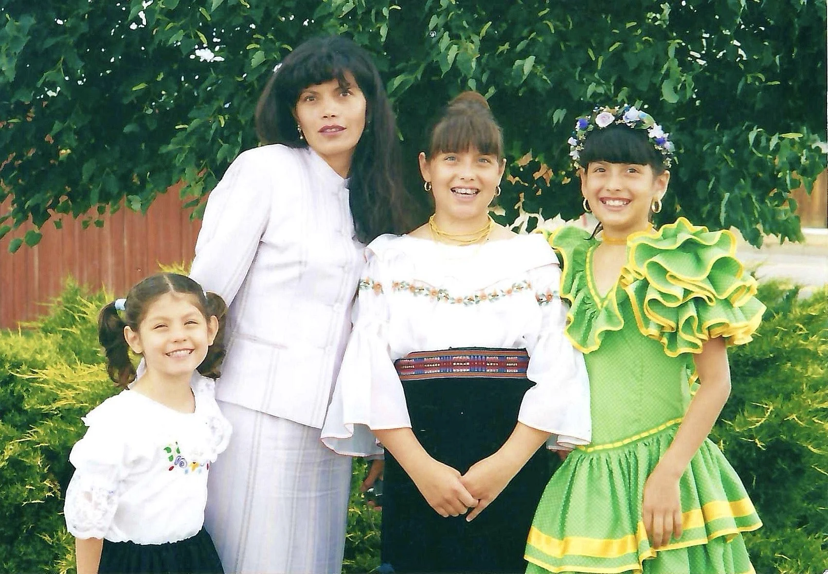 A group of five women standing outdoors in front of green bushes, dressed in traditional colorful Mexican dresses and white blouses, smiling at the camera.