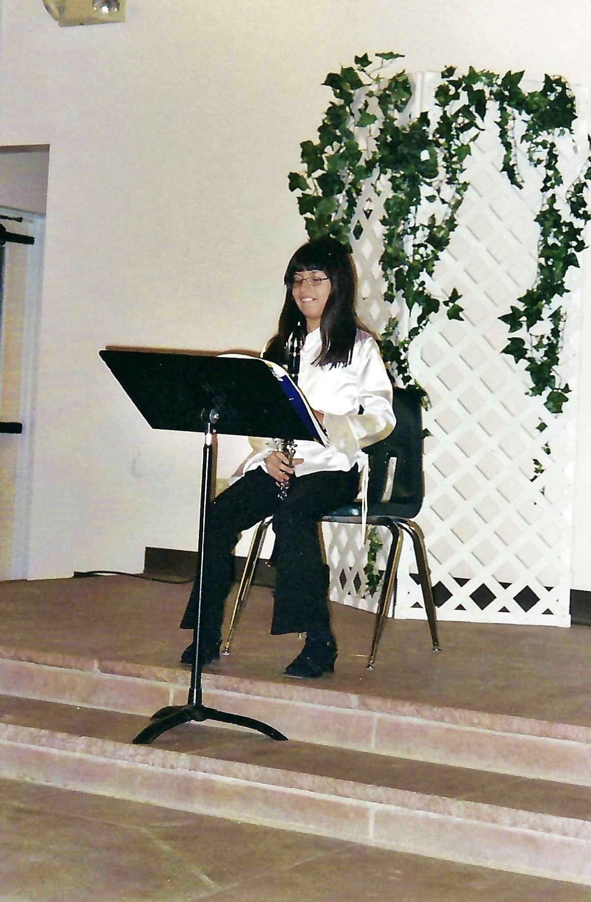 A woman sitting on a black chair on a small stage, holding a clarinet, with music stand in front of her. There is a decorative white lattice with green ivy behind her.