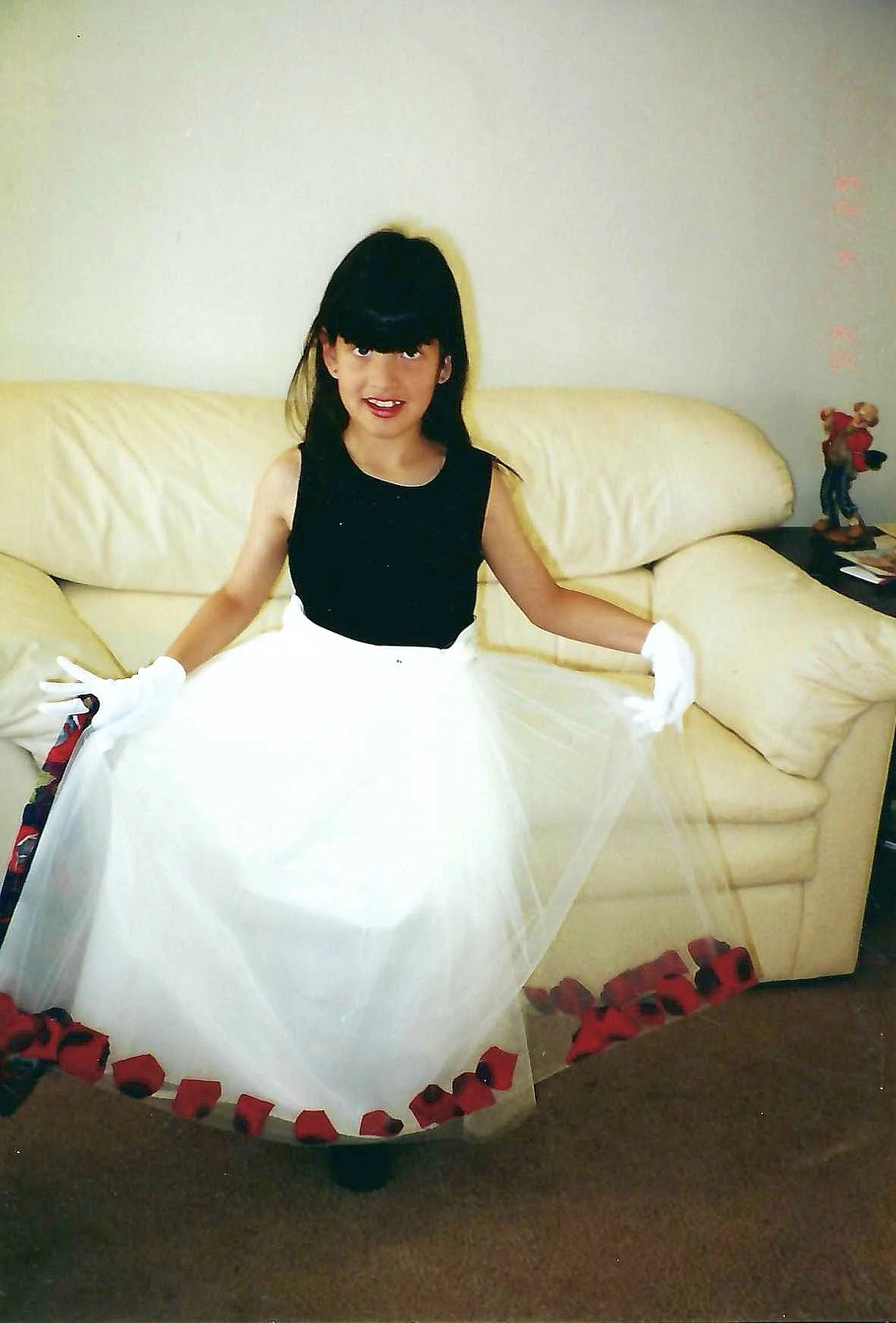 A woman dressed in a black top and white full skirt sitting on a yellow sofa, holding the skirt out around her. She has dark hair with bangs and is wearing white gloves. The sofa has pillows, and there are toy blocks on the floor and a decorative fig