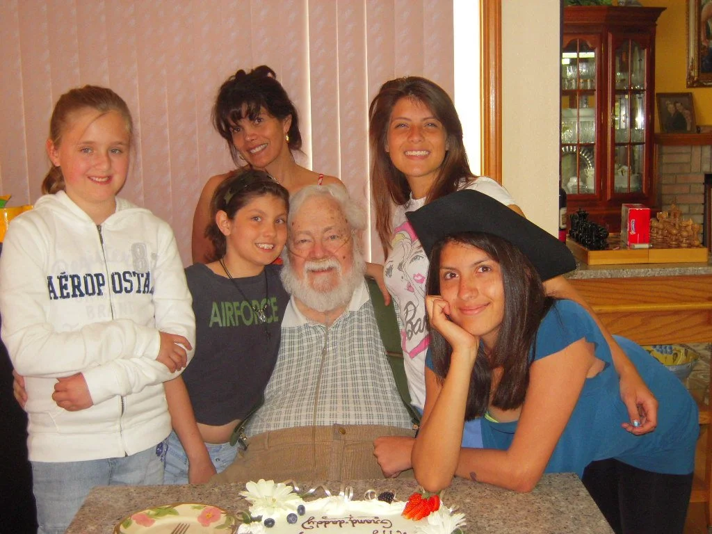 Group of six people, including children and adults, gathered around a birthday cake in a cozy kitchen or dining area, celebrating a special occasion.