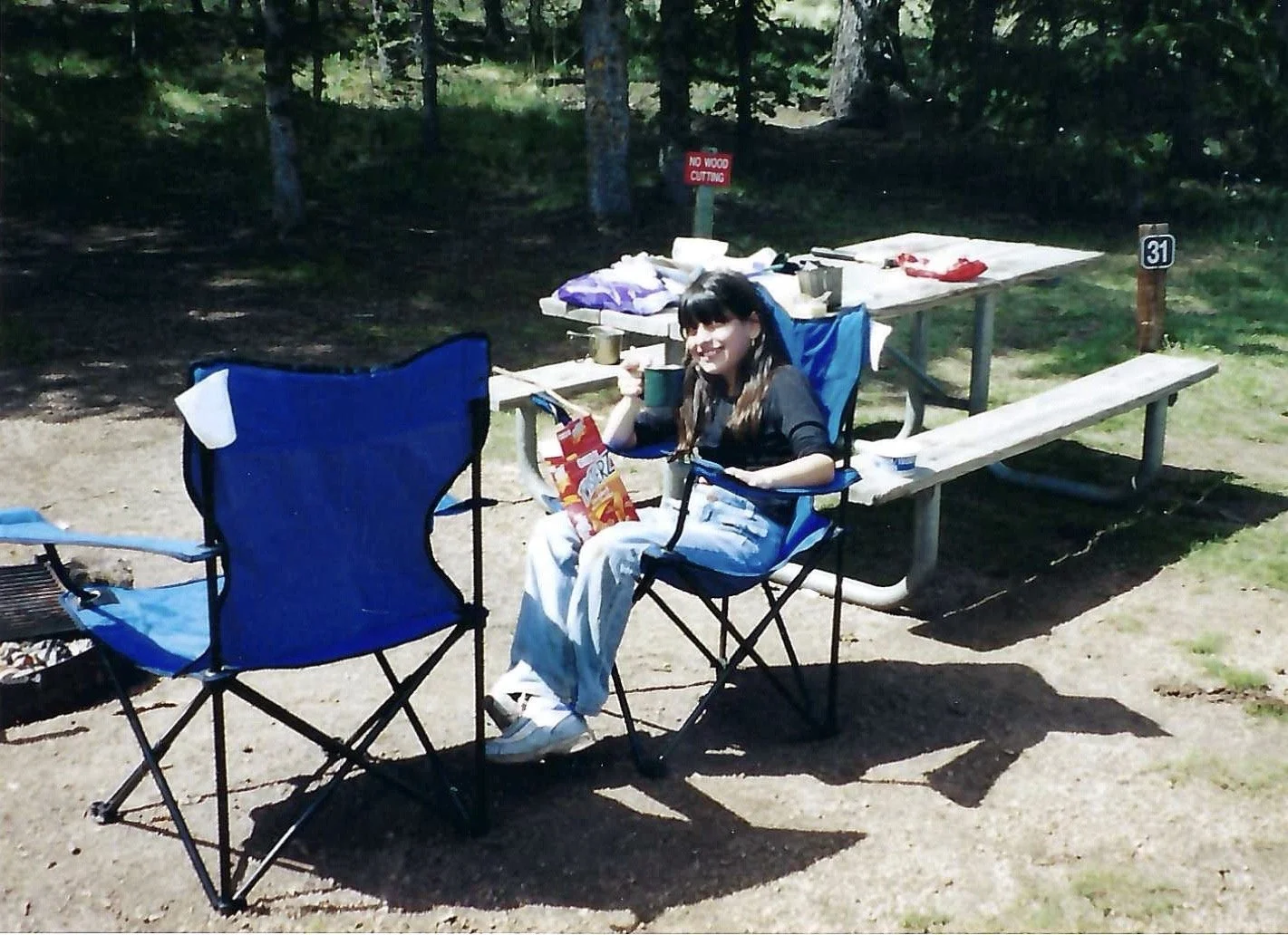 A young woman sitting in a blue camping chair at a picnic table in a wooded area, holding a mug and a bag of chips, with camping supplies and food on the table, under a sign that says 'No Wood Cutting'.