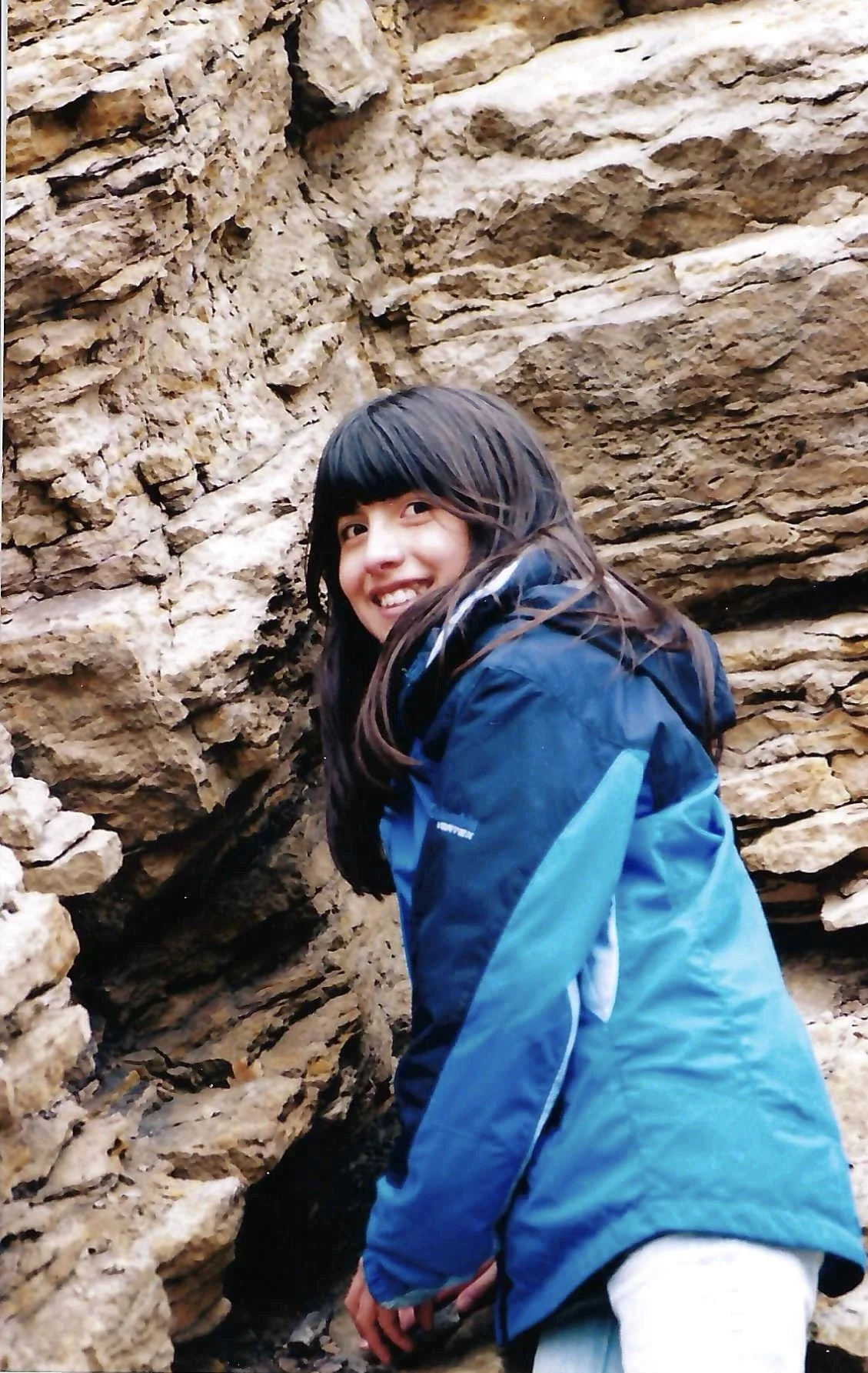 A girl with long black hair and a big smile, wearing a blue jacket, standing among layered rocks or cliffs.