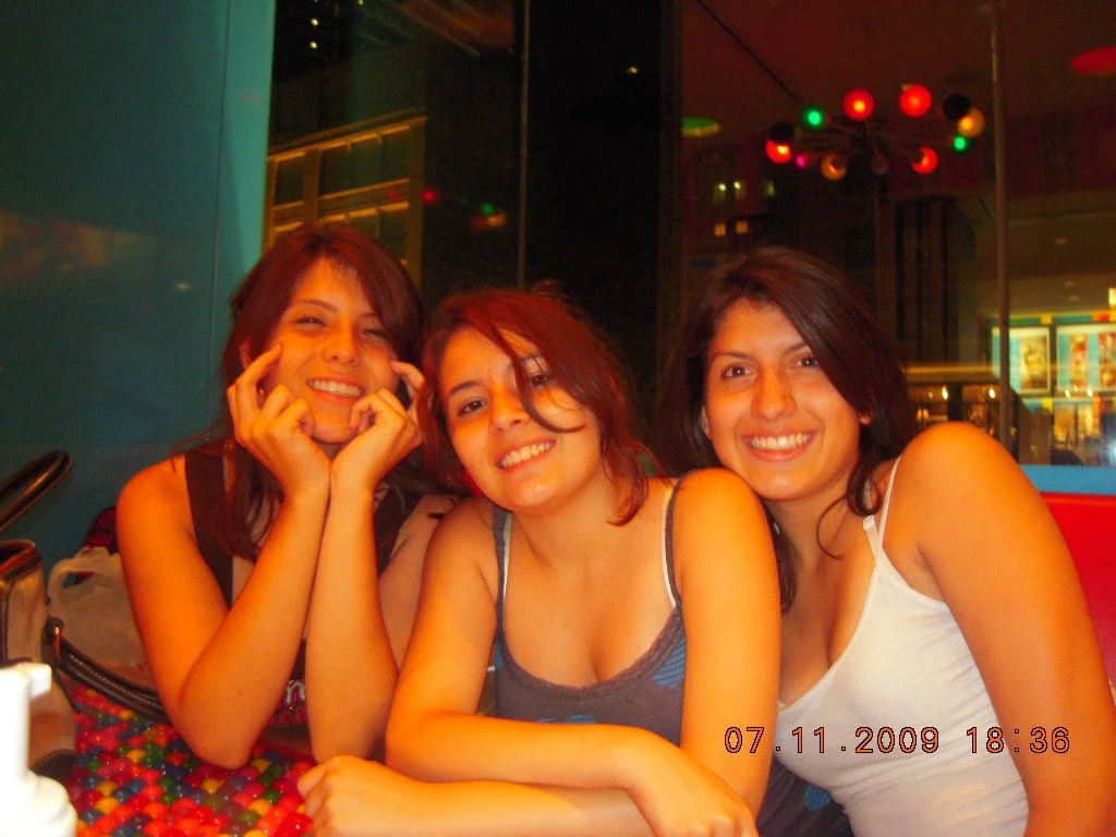 Three smiling young women sitting together in a restaurant at night, with colorful lights overhead and a reflection of a cityscape in the background.