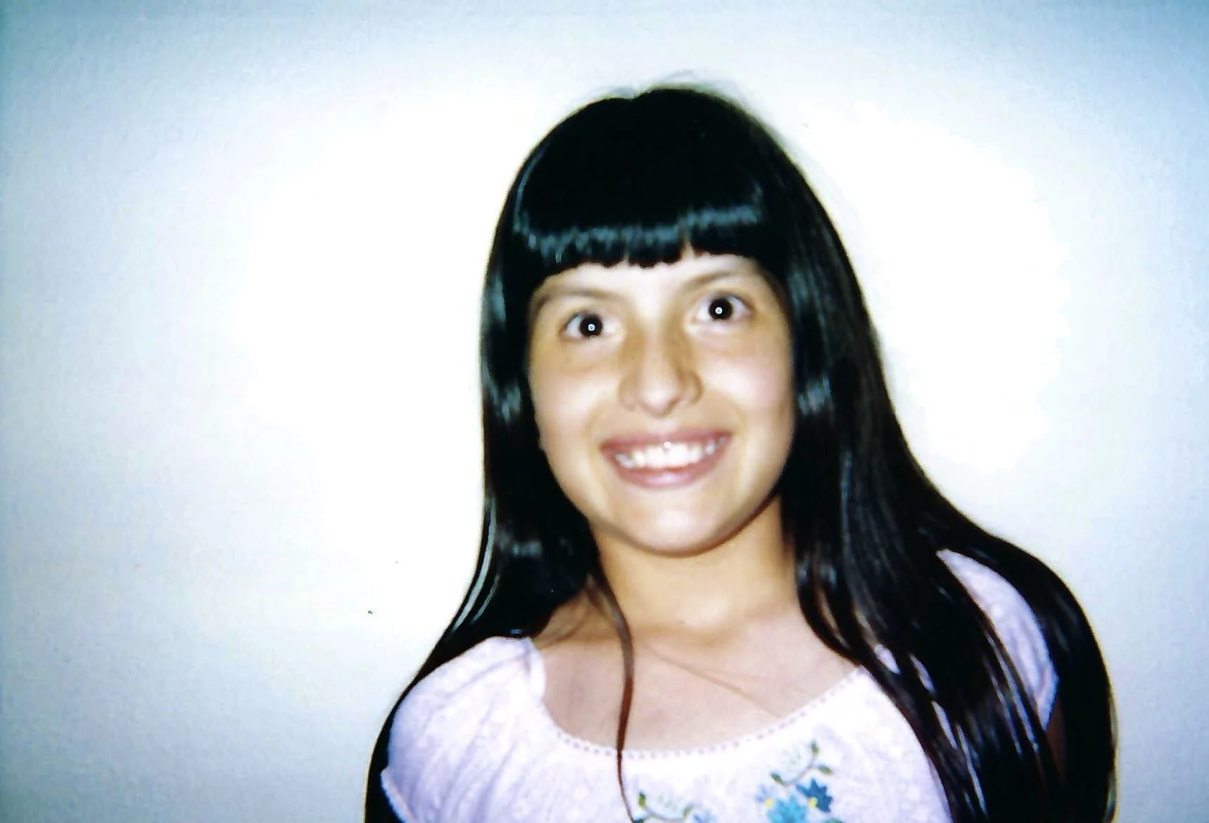 A young girl with long dark hair, light skin, and a bright smile, looking upward, wearing a light-colored top with floral embroidery on the front.