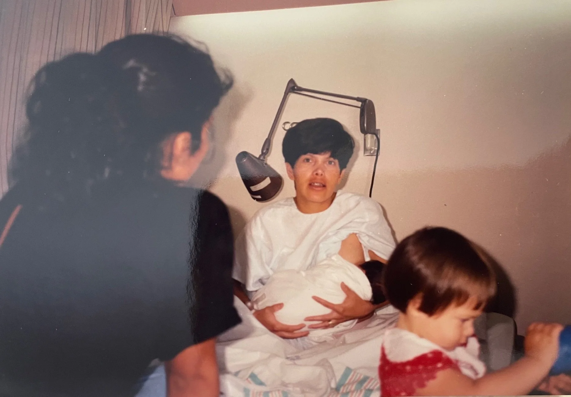 A woman in a hospital bed with short dark hair, wearing a white hospital gown, being attended to by multiple healthcare providers. One person is holding her arms, while another is placing a newborn baby in her lap. Several children are nearby, with o