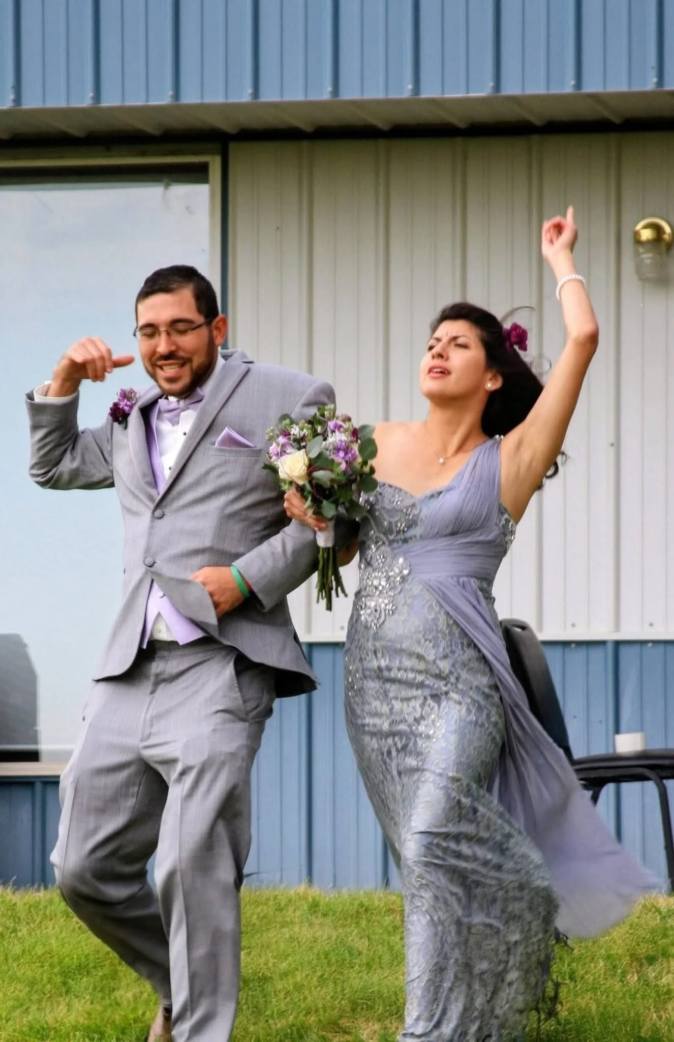 A man in a gray suit and a woman in a silver lace dress dancing outside on grass. The woman is holding a bouquet and has her arm raised. Both appear to be enjoying themselves at a celebration or wedding.