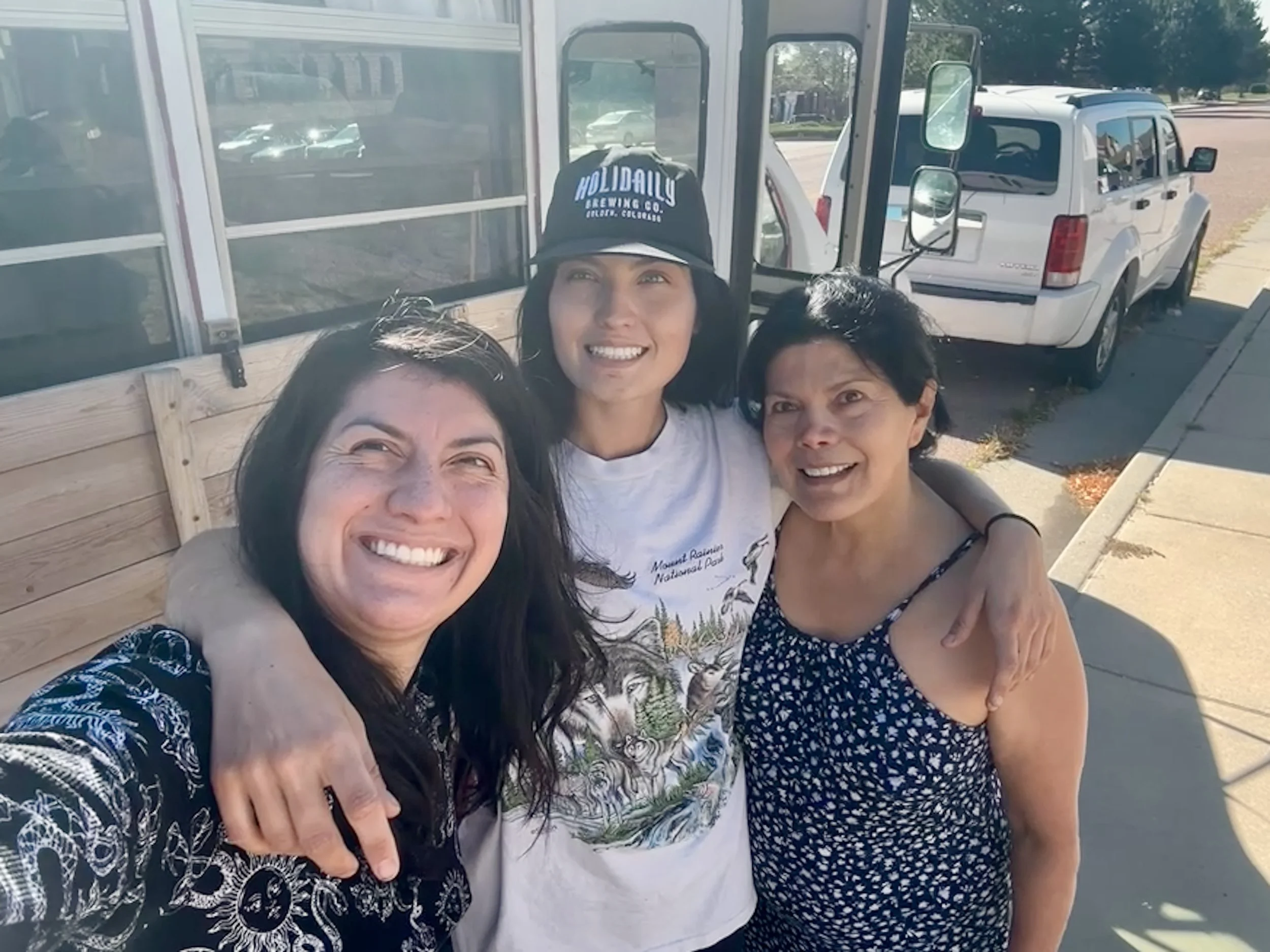 Four women smiling for a selfie outdoors near a white vehicle and a wooden structure, wearing casual summer clothing.