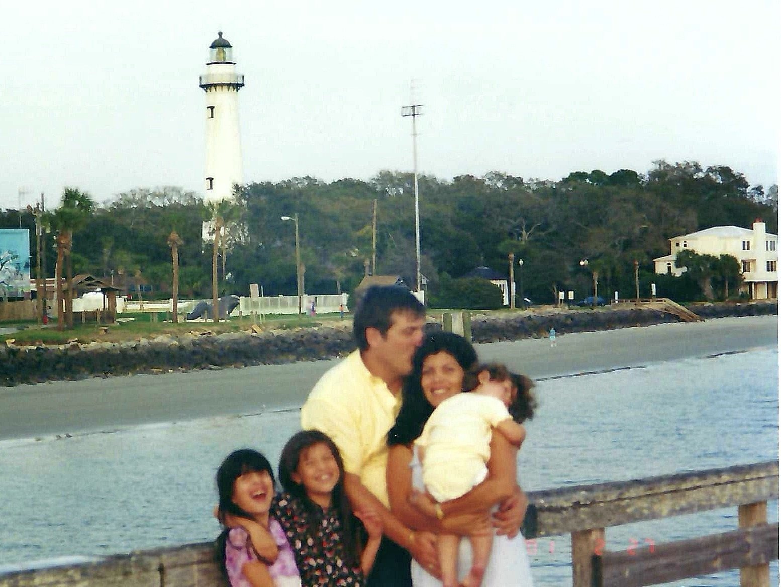 A family of five stands on a beach with water and a promenade behind them, featuring a lighthouse, trees, and buildings in the background.
