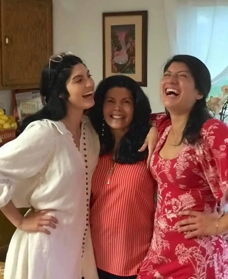 Three women smiling and laughing together in a cozy kitchen, dressed in casual and colorful clothing.
