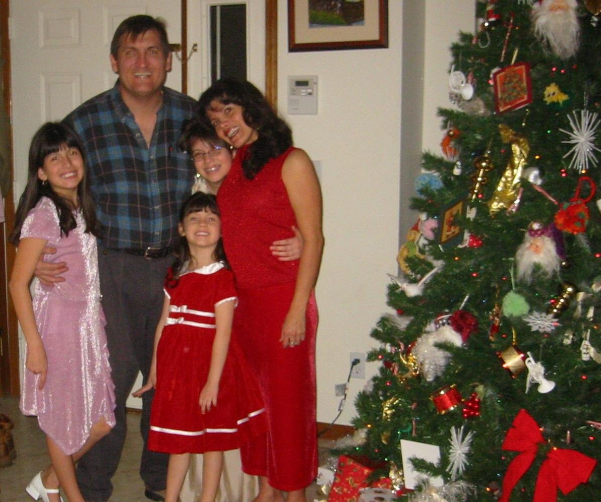 A family of five celebrating Christmas indoors, standing next to a decorated Christmas tree with presents underneath.