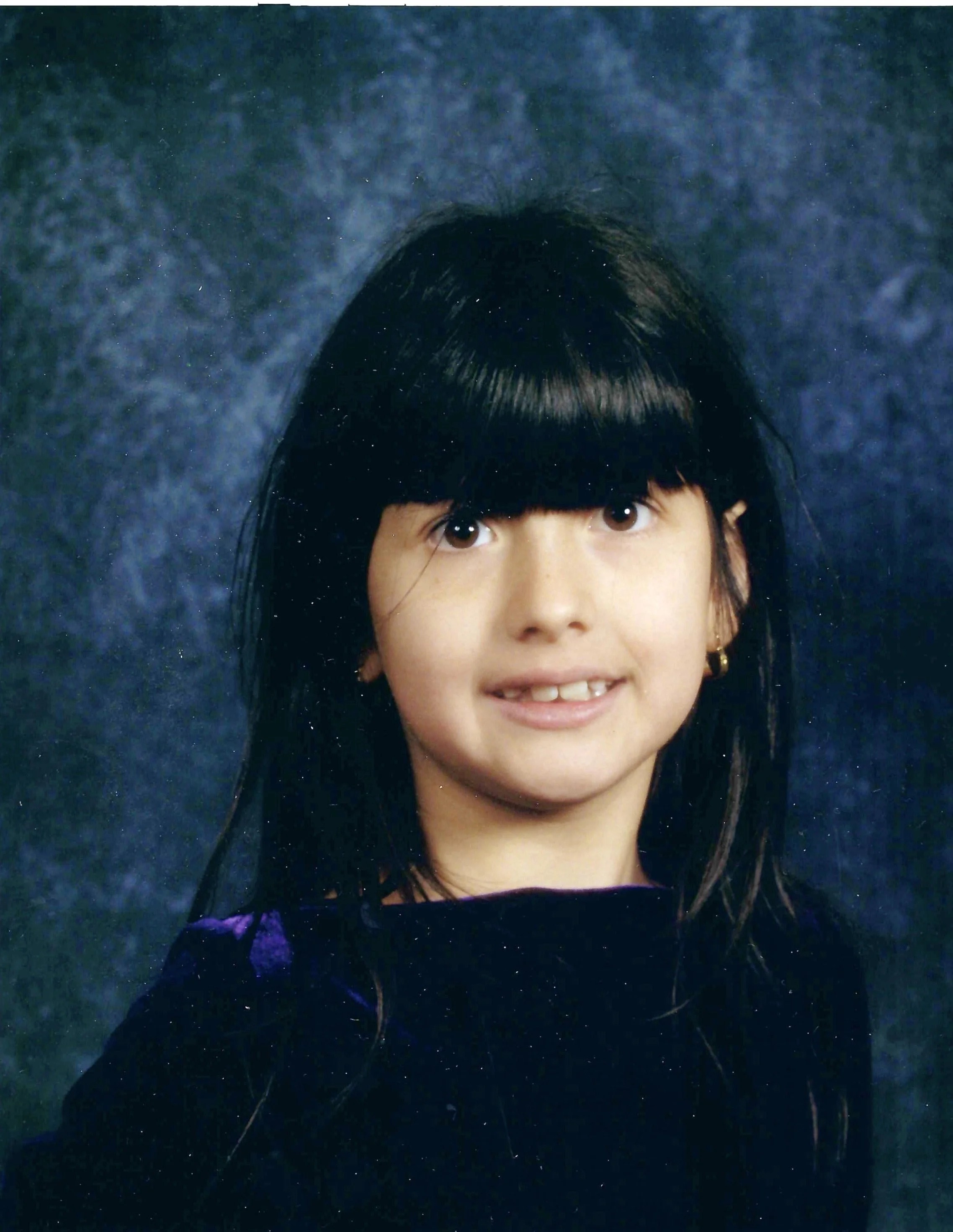 A young girl with black hair and brown eyes smiling at a camera, against a dark, starry background.