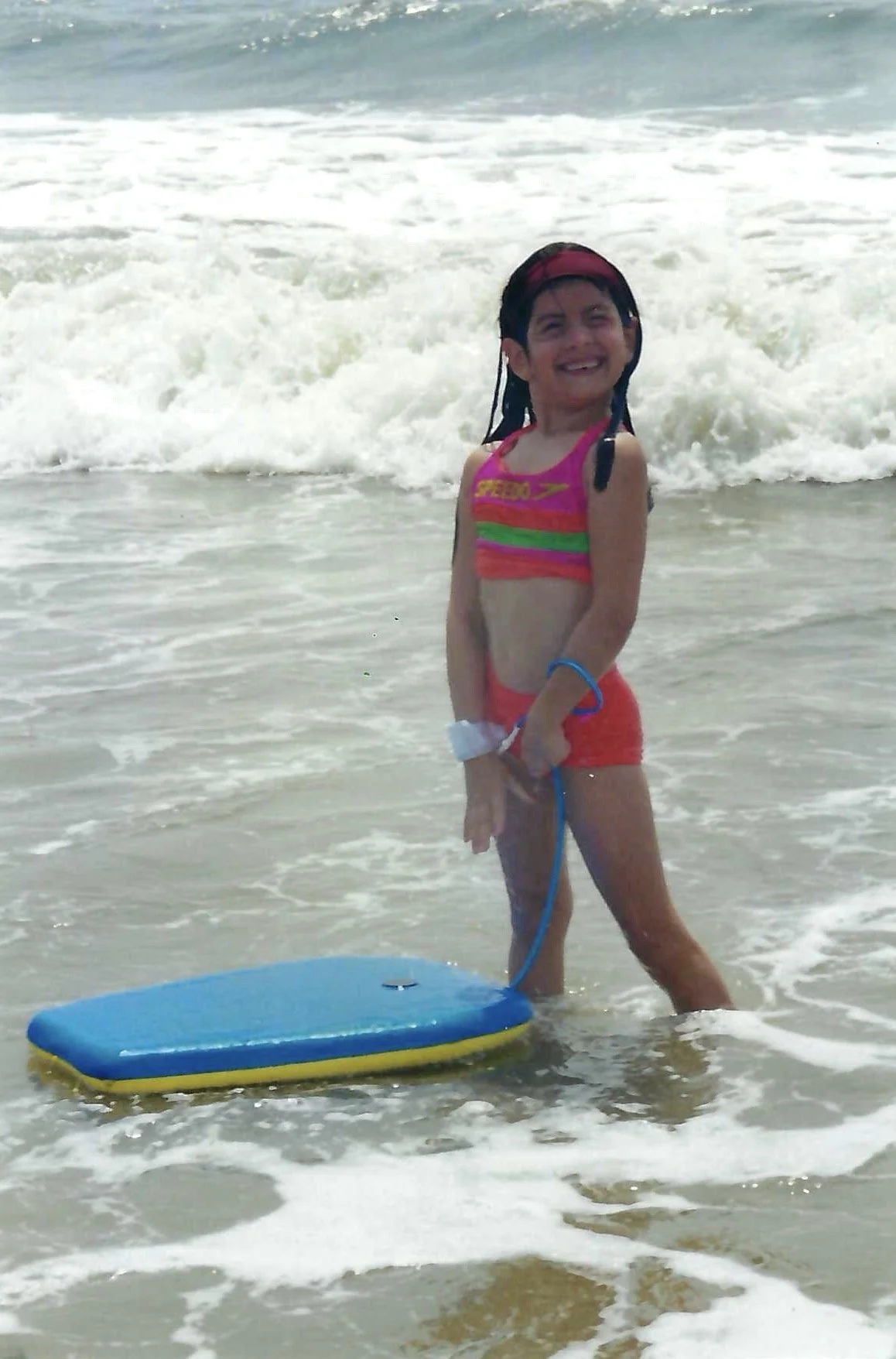 A young girl in a pink and green swimsuit standing in shallow ocean water, smiling, holding a blue and yellow float board, with waves in the background.