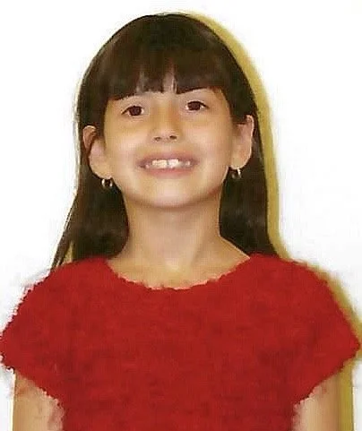 Smiling young girl with dark hair, wearing a red top and small hoop earrings.