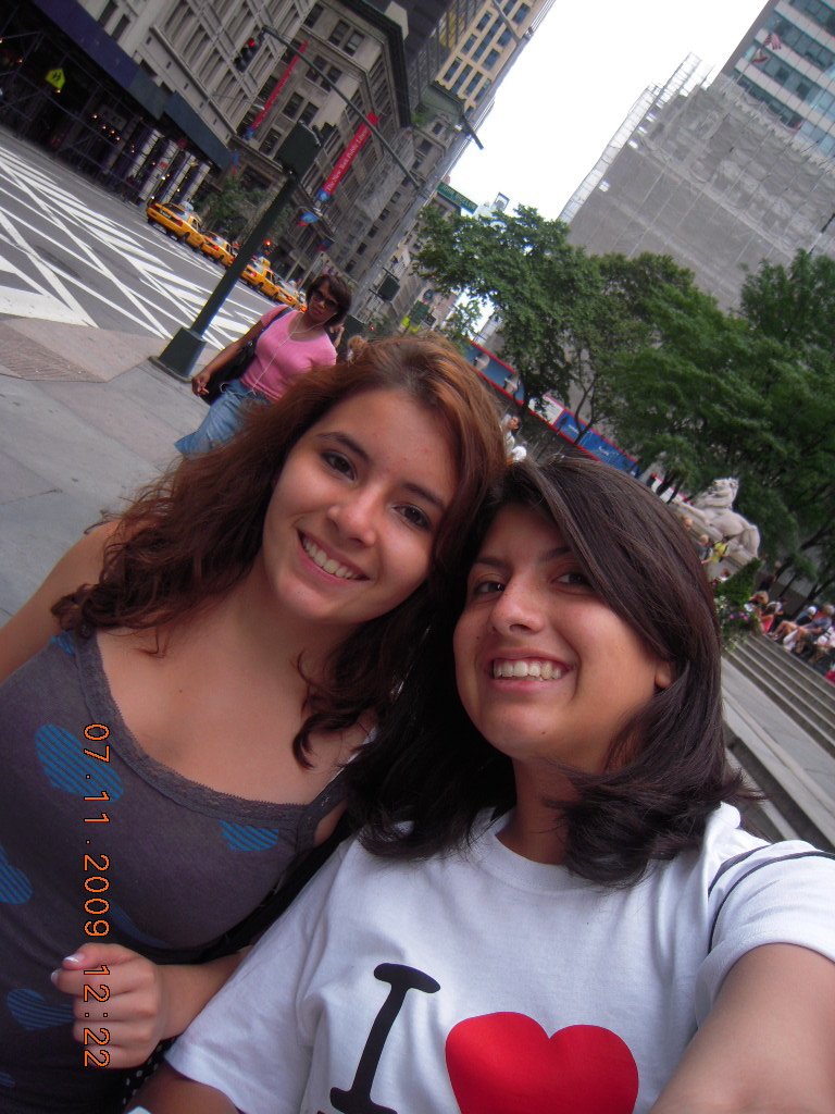 Two women taking a selfie on a city sidewalk with tall buildings and trees in the background. One woman has dark brown shoulder-length hair and is wearing a white shirt with a red heart, and the other has curly reddish-brown hair and is wearing a gra