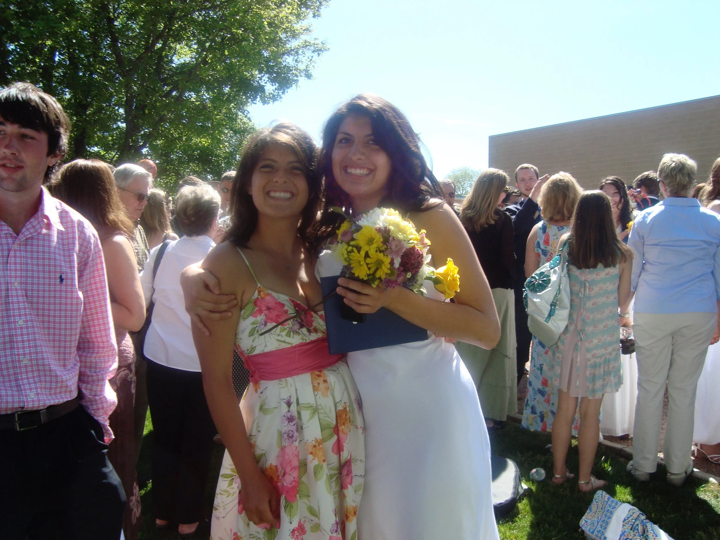 Two women smiling, one holding a bouquet of flowers, at an outdoor gathering under a tree on a sunny day with many people in the background.