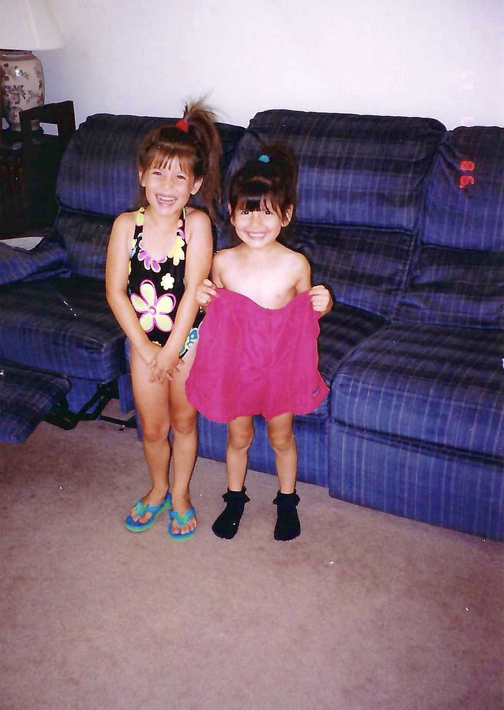 Two young girls standing together in a living room, smiling. One girl is wearing a colorful swimsuit with a flower pattern and flip-flops, and the other girl is holding a pink towel, wearing black socks.