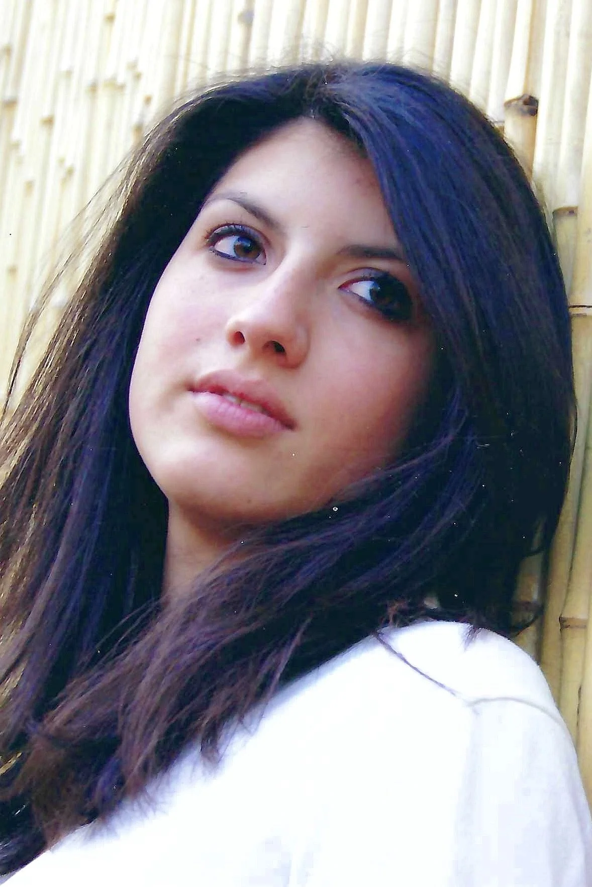 Close-up of a young woman with long dark hair, standing in front of a bamboo fence.