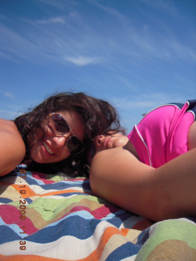 Two women lying on a colorful striped blanket outdoors on a sunny day. One woman is smiling and wearing sunglasses, while the other has her face turned away and is resting her head on her arm. The sky is clear and blue.