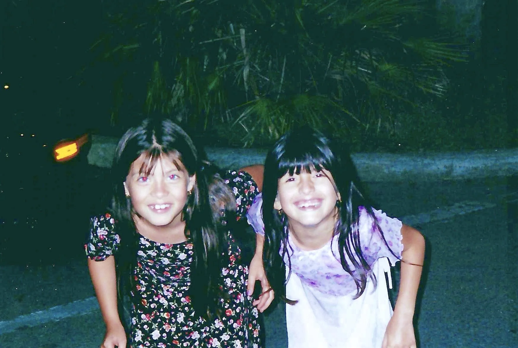 Two young girls with dark hair, wearing colorful dresses, smiling and standing outdoors at night in front of green plants.