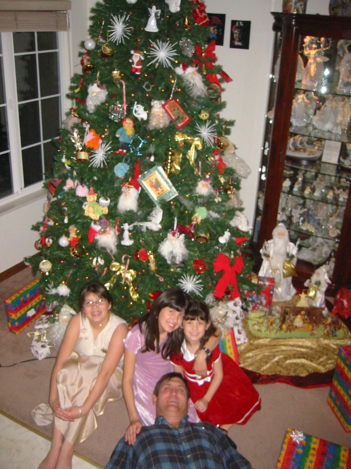A Christmas tree decorated with ornaments, ribbons, and lights, with four children and one adult posing in front of it, inside a home with a window and a display cabinet.