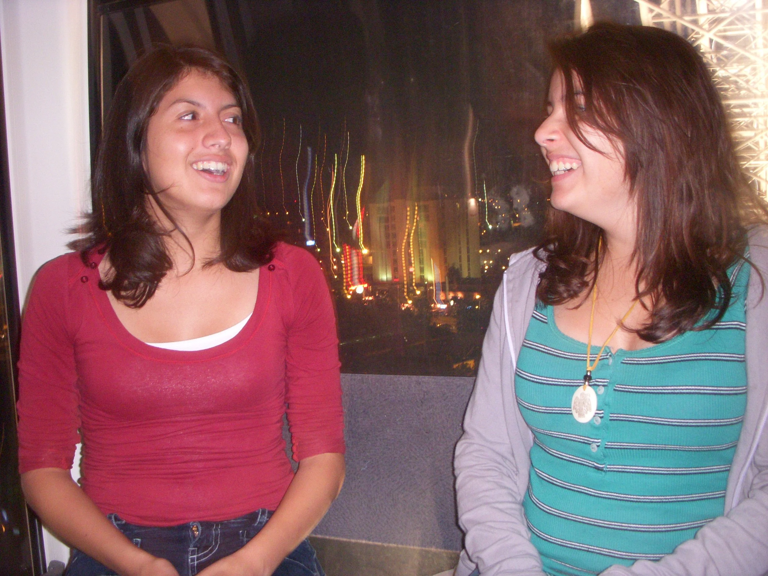 Two women with dark brown hair smiling and talking, indoors at night with city lights visible through the window behind them.