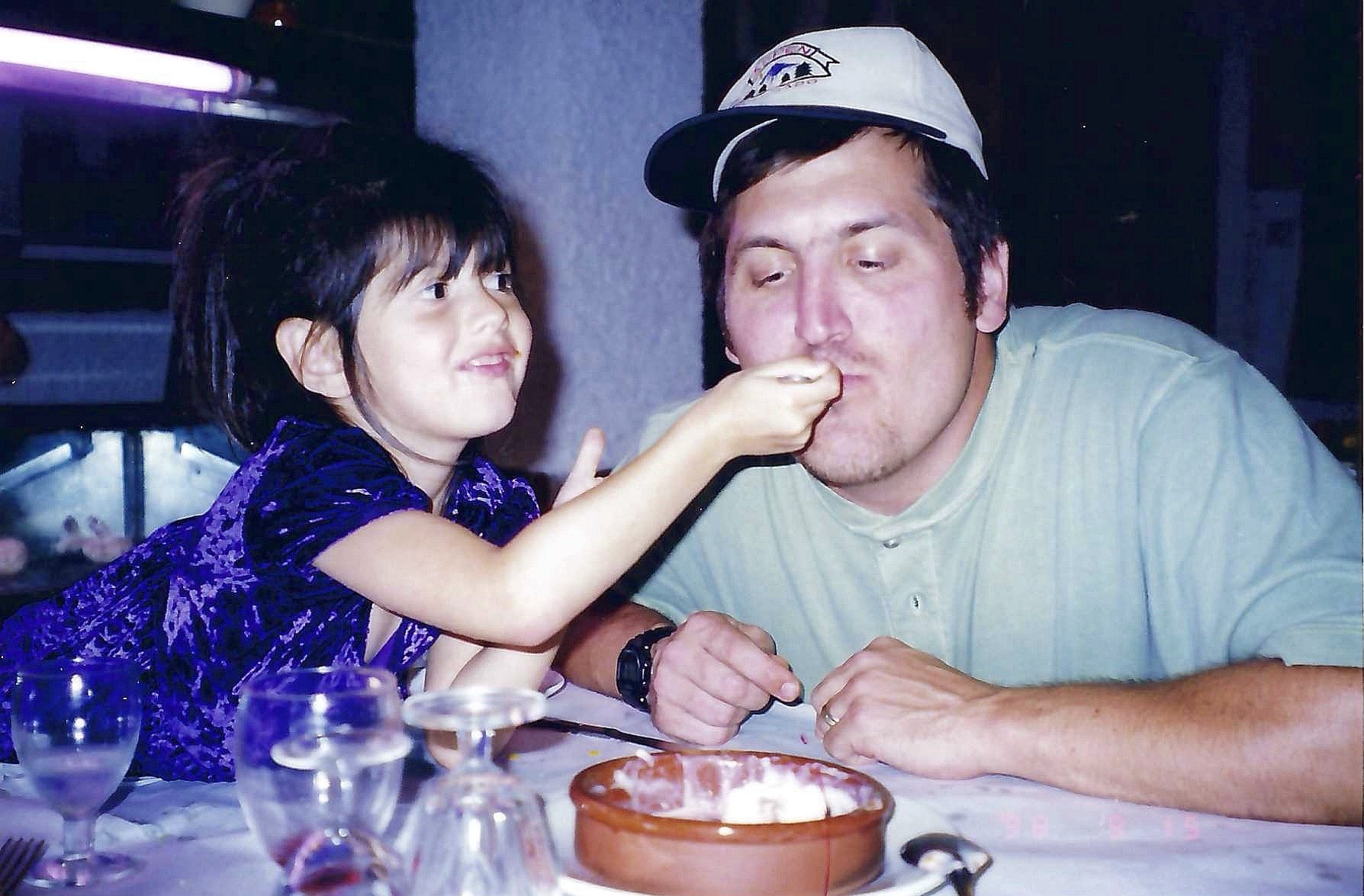 A young girl with dark, curly hair wearing a purple dress feeds a man with brown hair and a beard, who is wearing a light-colored shirt and a baseball cap, at a restaurant table. There is food and drink on the table.