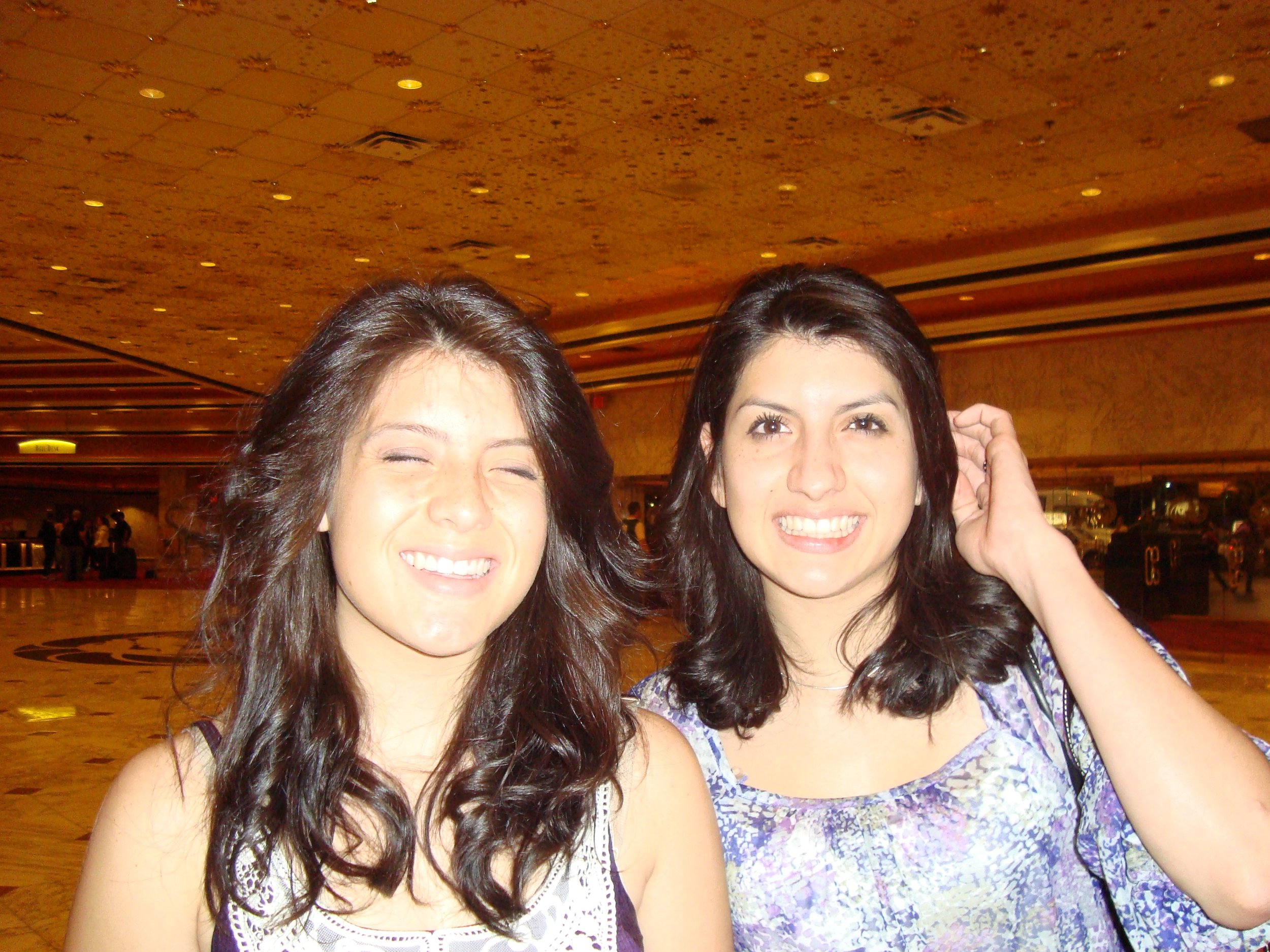 Two women with dark brown hair smiling in an indoor setting with a gold-colored ceiling and warm lighting.
