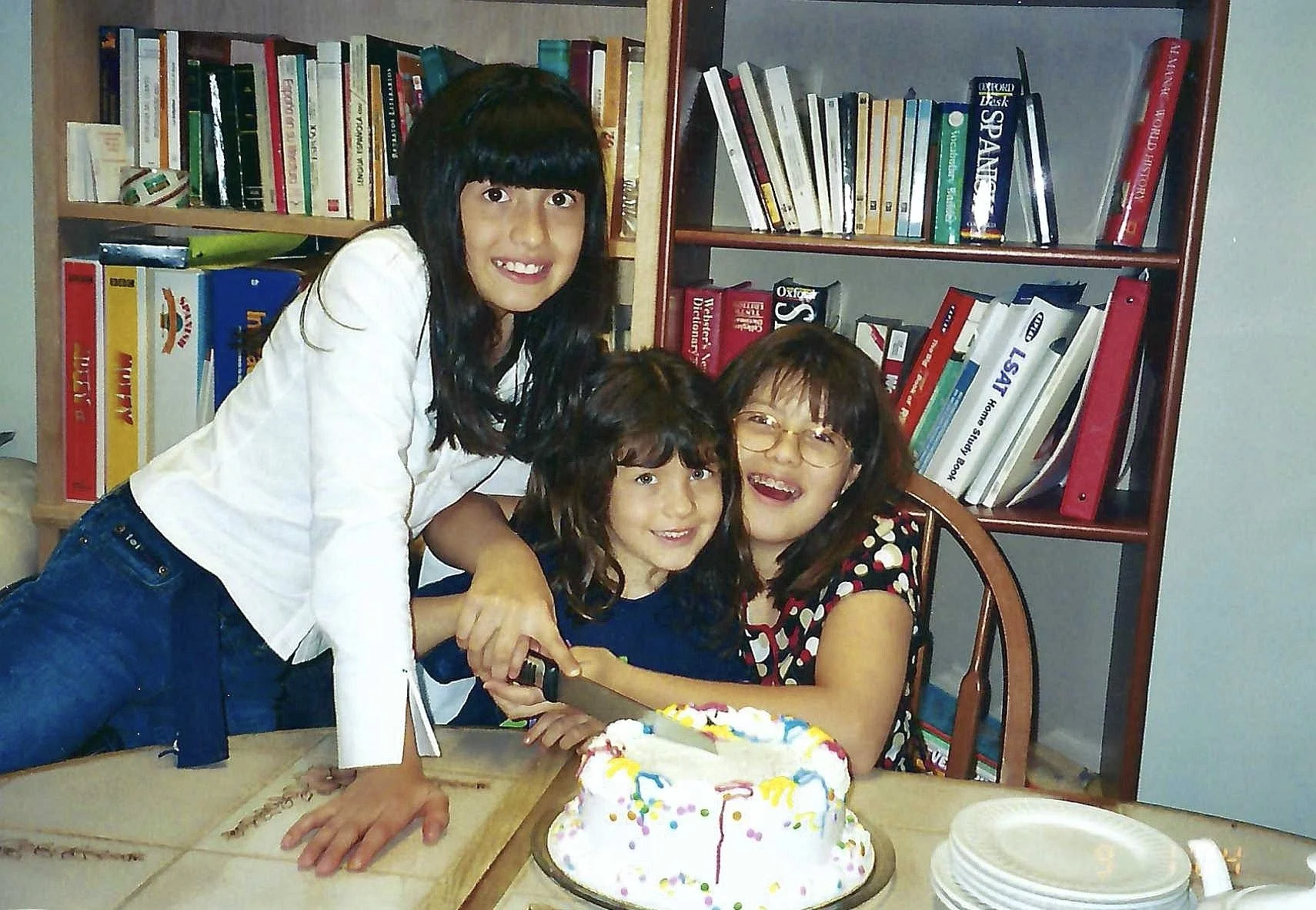 Three females are gathered around a table celebrating a birthday. A girl with long dark hair and a white shirt stands next to two seated females, a woman with glasses and a girl with curly hair, both smiling. There are bookshelves in the background, 