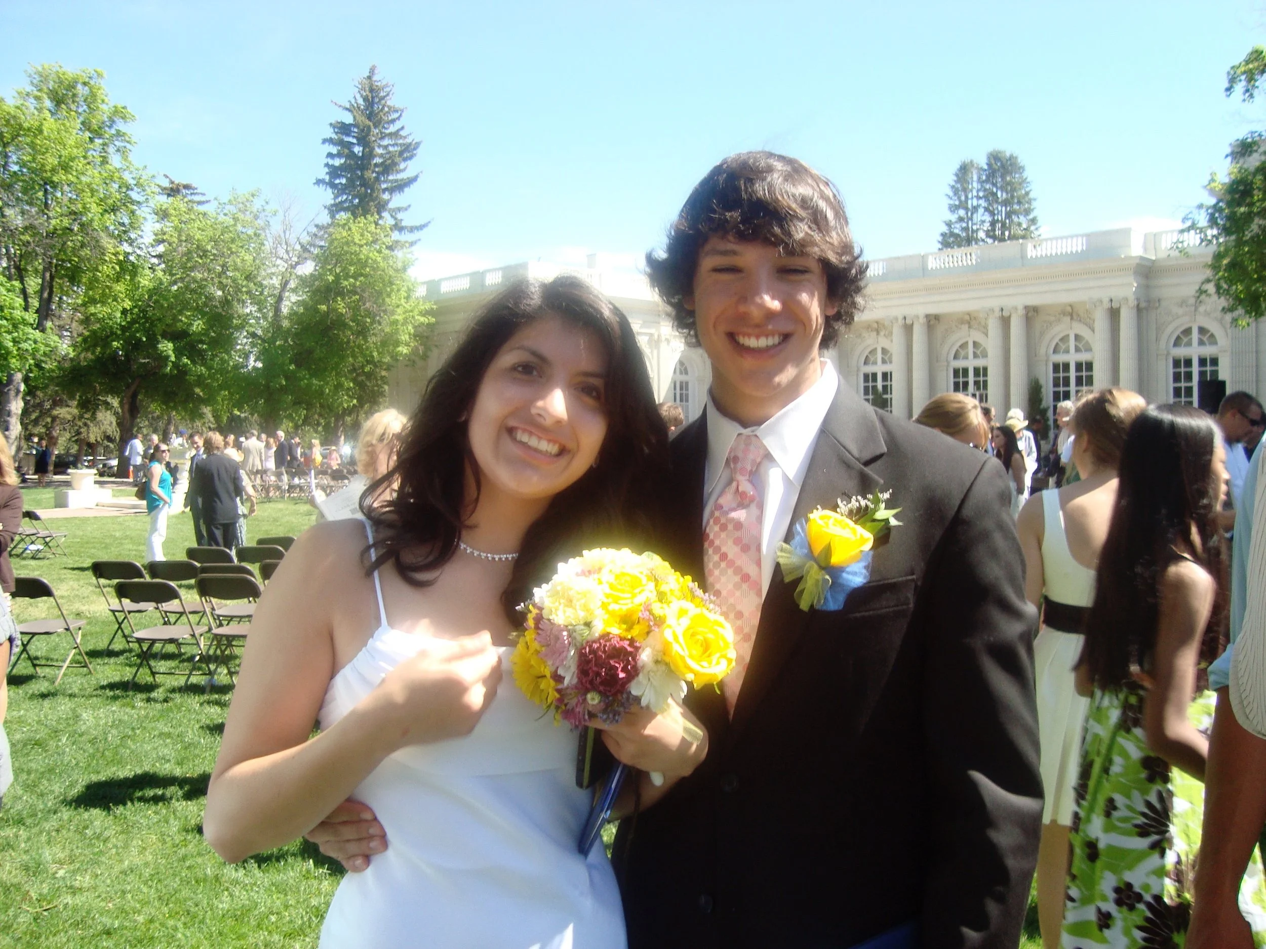 A wedding couple smiling outdoors, with the bride holding a bouquet of yellow, pink, and white flowers, and the groom wearing a boutonniere, during a sunny day with many guests in the background and classical architecture in the distance.