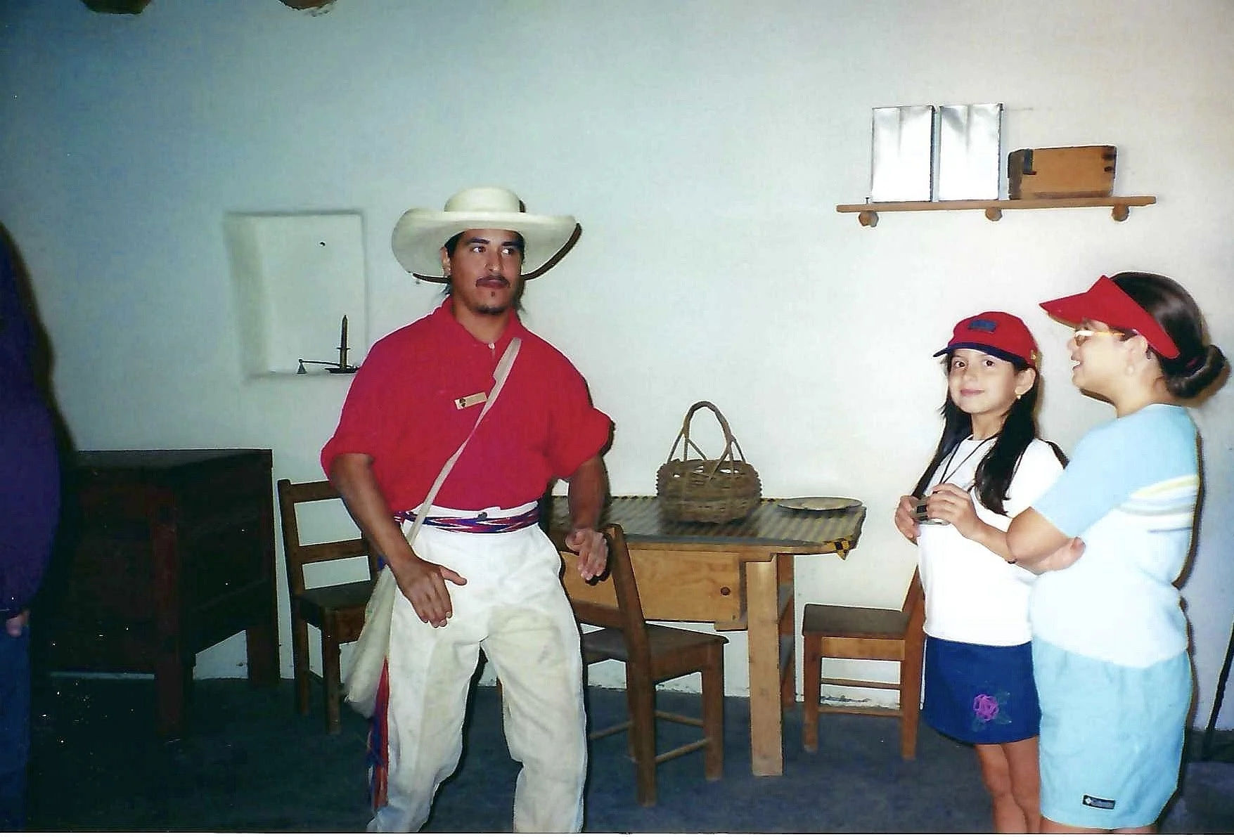 A man wearing traditional Mexican attire, including a wide-brimmed hat and red shirt, is standing in a room with two young girls. One girl has a red visor and is holding a small container, while the other girl wears glasses and a red visor as well. T