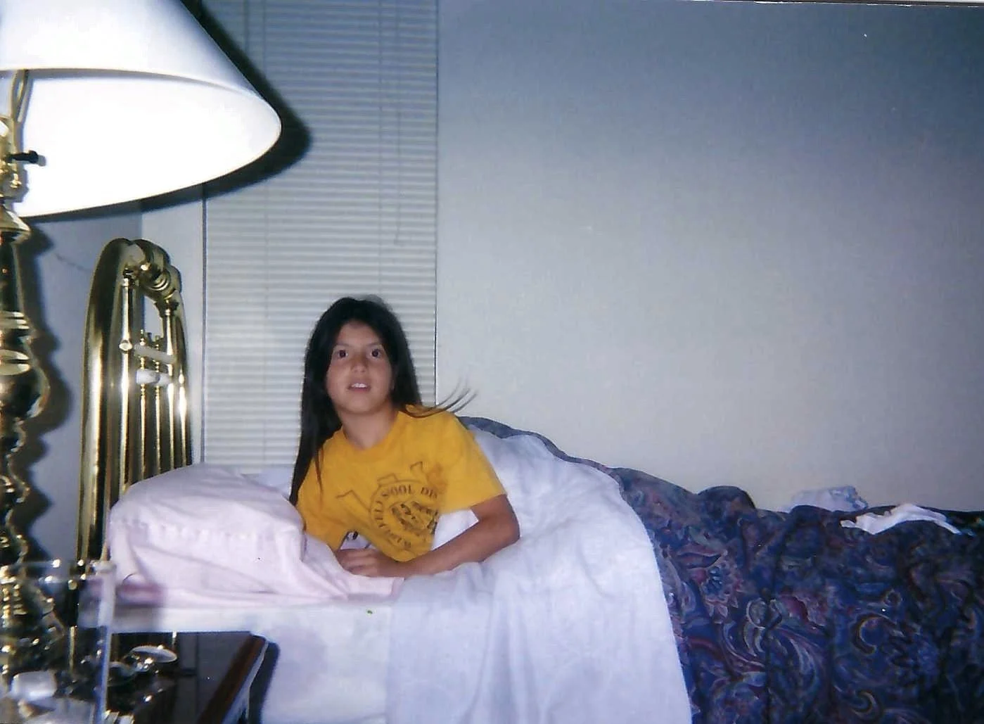 A young woman with long dark hair laying in bed, wearing a yellow T-shirt with a logo, in a dimly lit room with a brass bed frame and a gold-colored lamp.