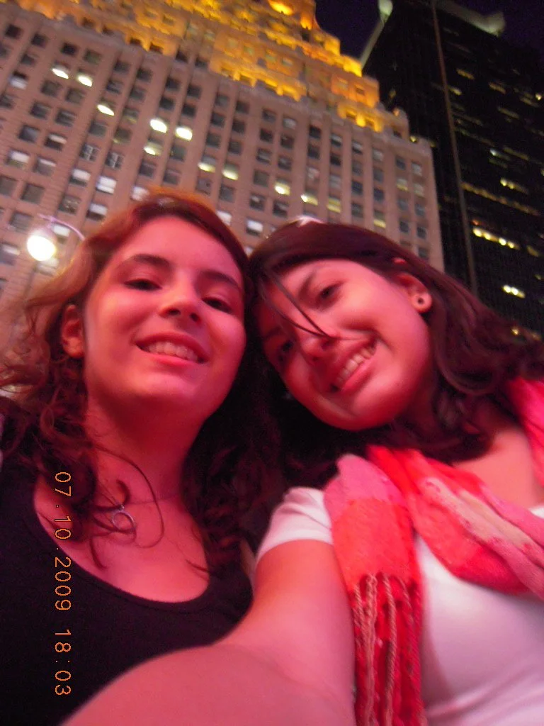 Two women smiling for a selfie in front of a tall, illuminated building at night.