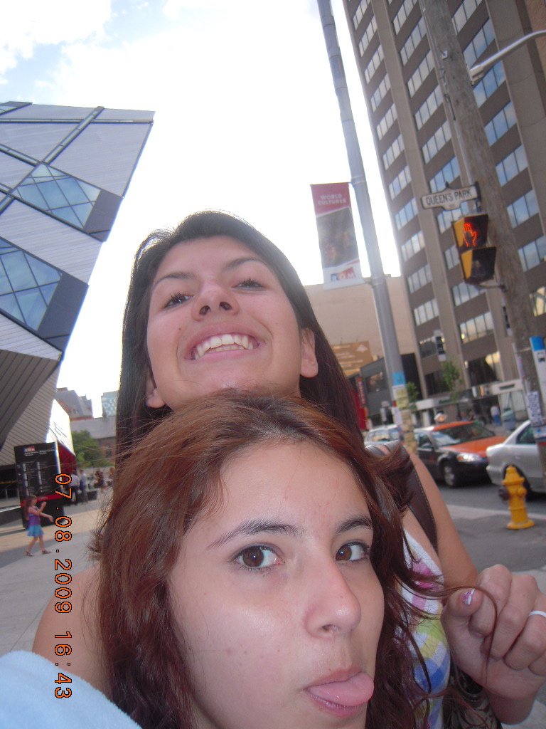 Two young women taking a selfie outdoors on a sunny day in an urban area with tall buildings and cars in the background.