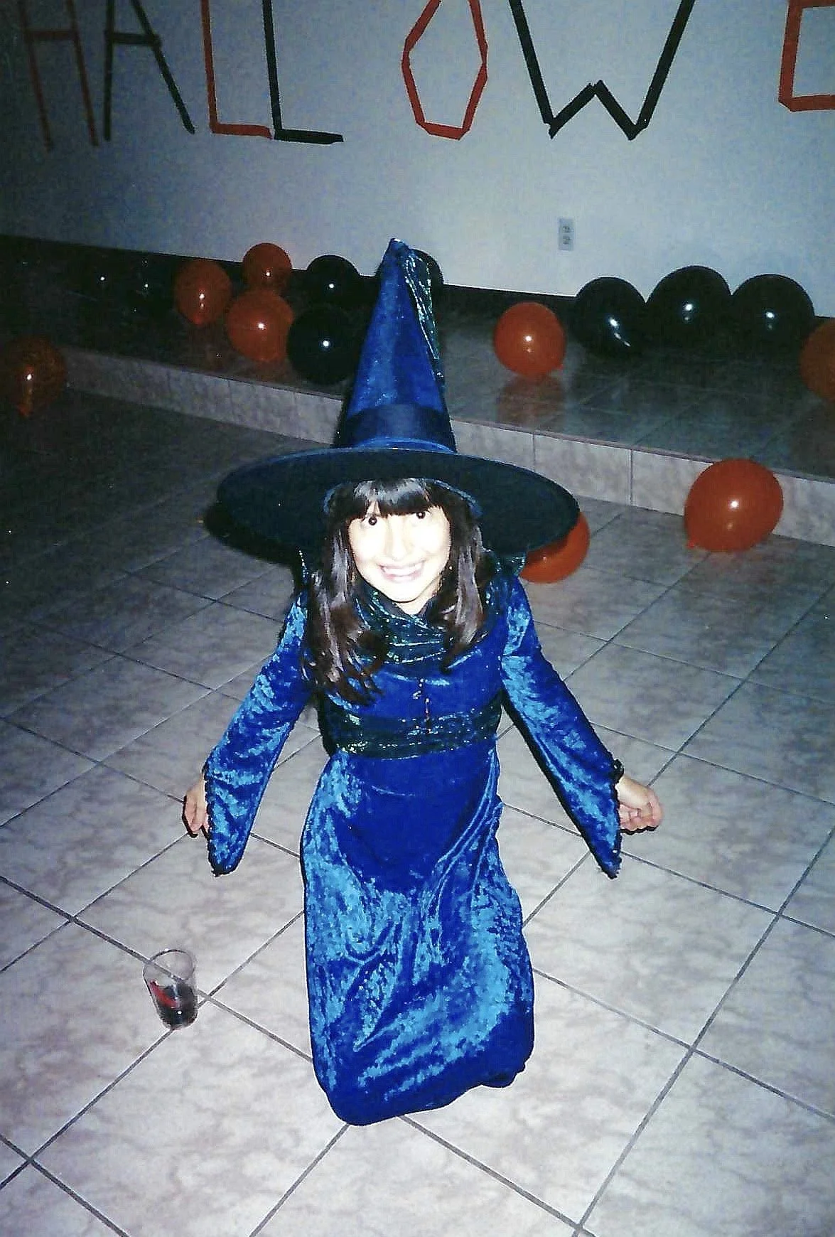 A young girl dressed as a witch in a blue velvet gown and a pointy hat, standing on a tiled floor with balloons and a banner that reads 'HALLOWEEN' in the background.