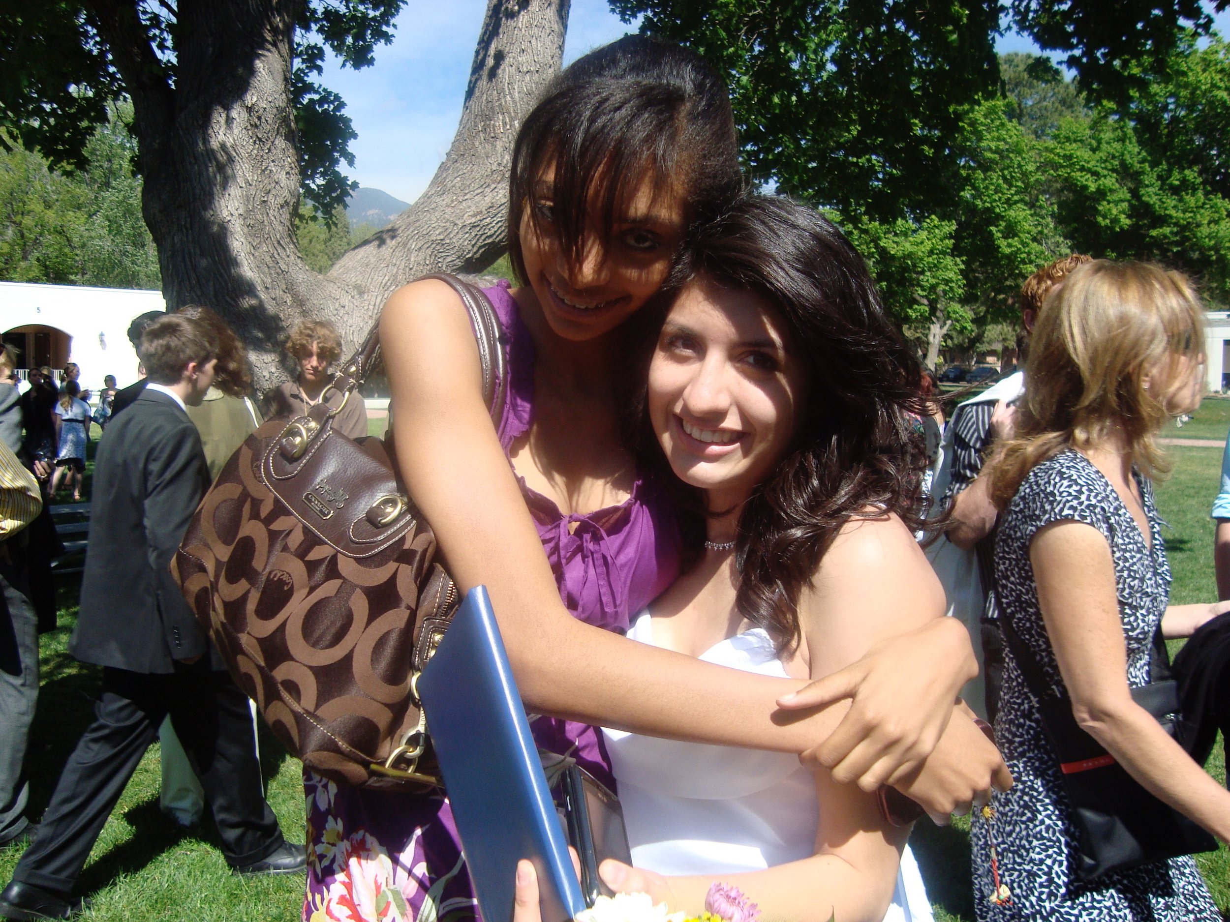 Two women hugging and smiling at an outdoor event, with multiple people and trees in the background.