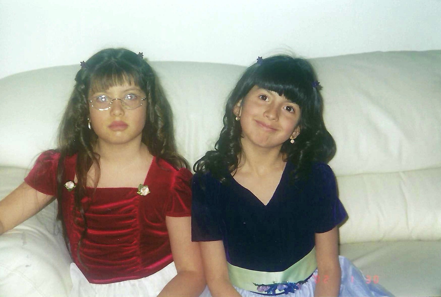 Two young girls sitting on a white couch, one wearing glasses and a red top and the other in a black top and colorful skirt, both smiling.