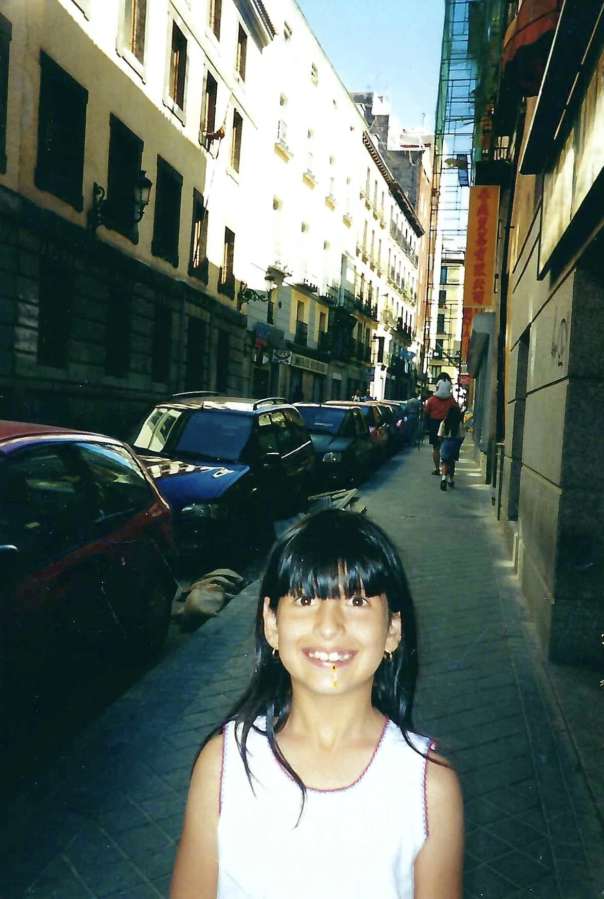 A young girl with black hair and bangs smiling on a city sidewalk with parked cars and buildings in the background.