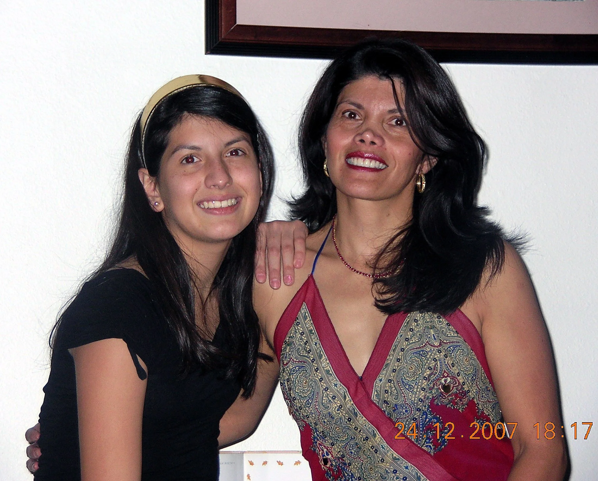 Two women standing side by side, smiling at the camera, in front of a white wall with a framed picture. One woman is wearing a black dress with cutout sleeves and has long dark hair, while the other is wearing a colorful, ornate dress with a deep nec