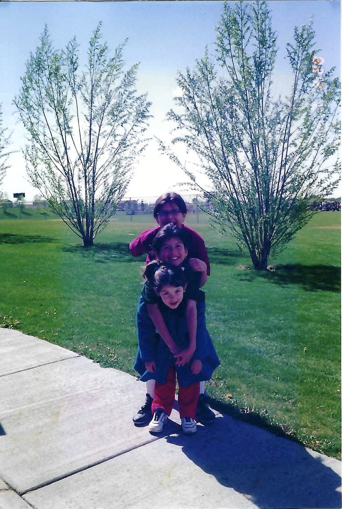 Three children playing outdoors on a sidewalk with green grass, trees, and blue sky in the background. The oldest child is standing behind the other two, with the youngest children in front, one of whom is carrying the other on their back.