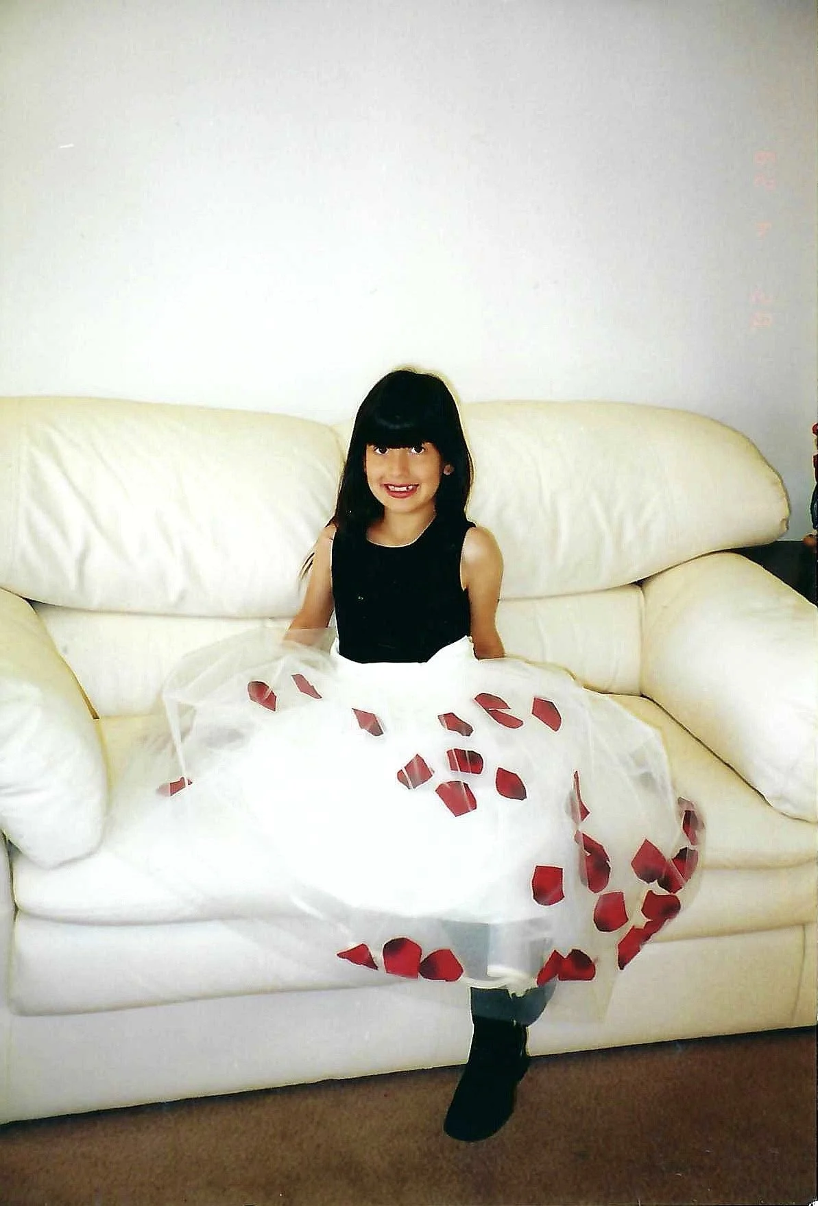 A young girl with dark hair and a black dress sitting on a white couch, holding a white dress with red heart-shaped patterns.