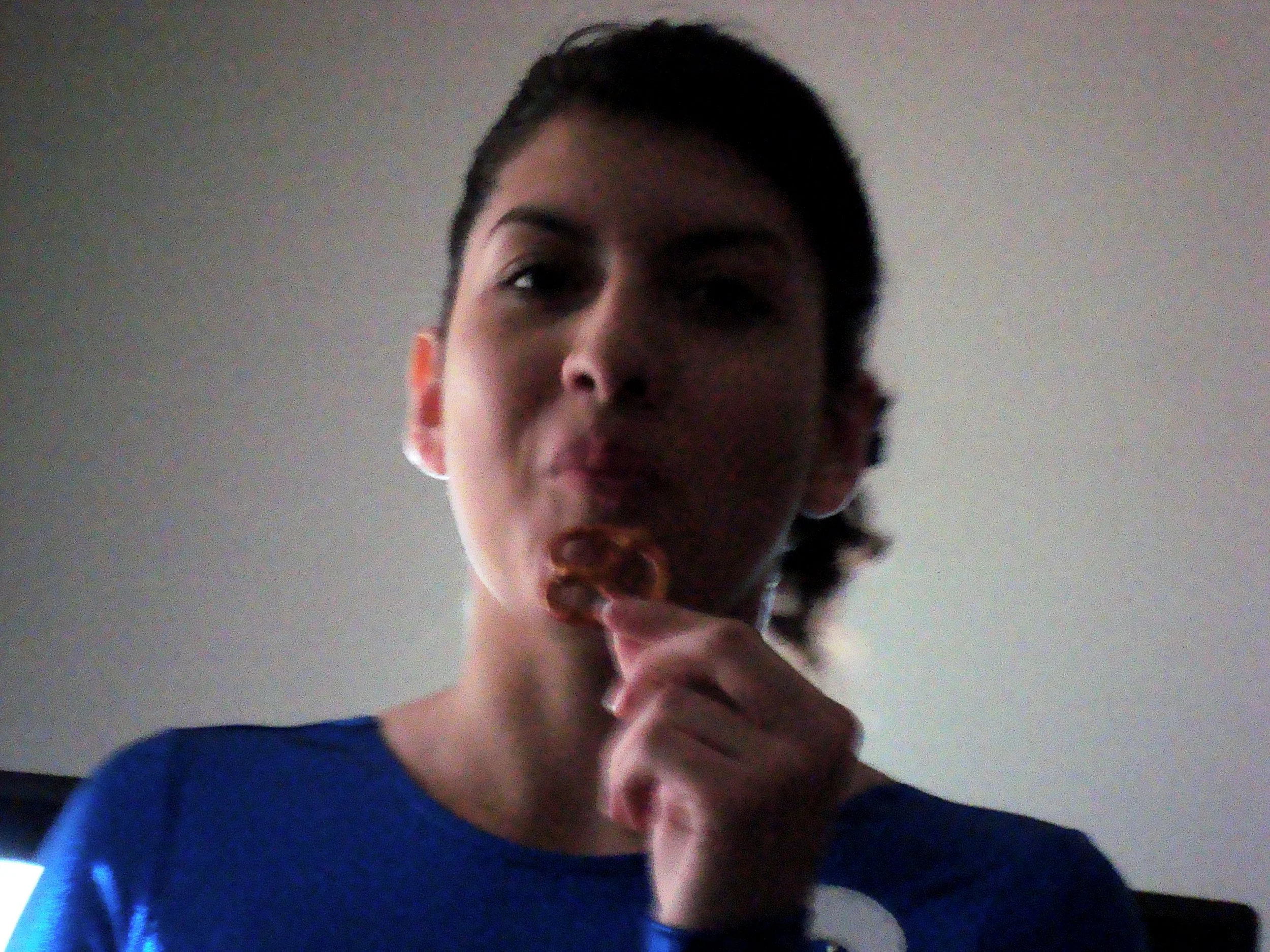 Young person with short dark hair making a kissy face, wearing a blue shirt and resting their chin on their hand, against a plain wall background.