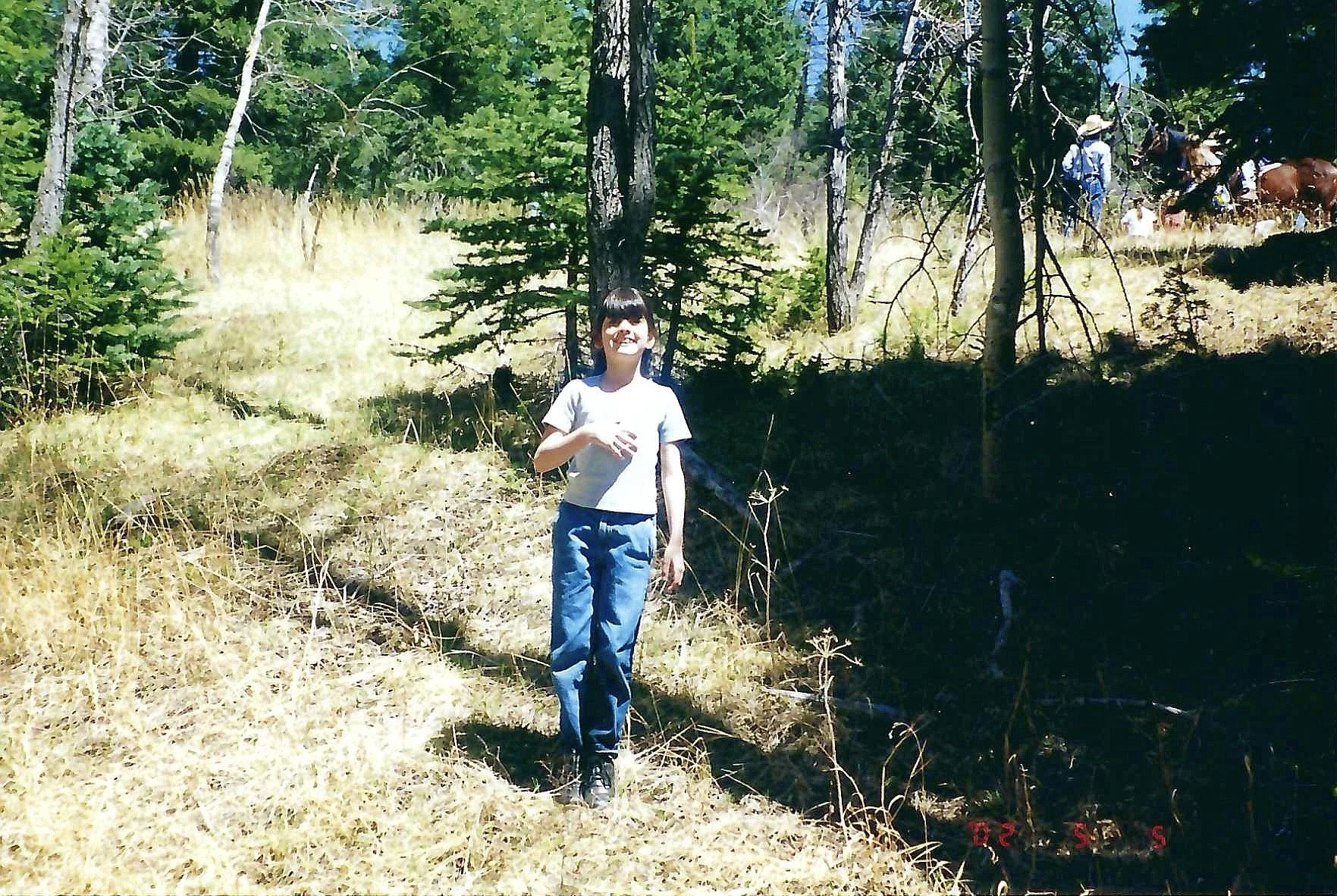 A girl standing in a wooded area on a grassy hill, with trees and a person on horseback in the background.