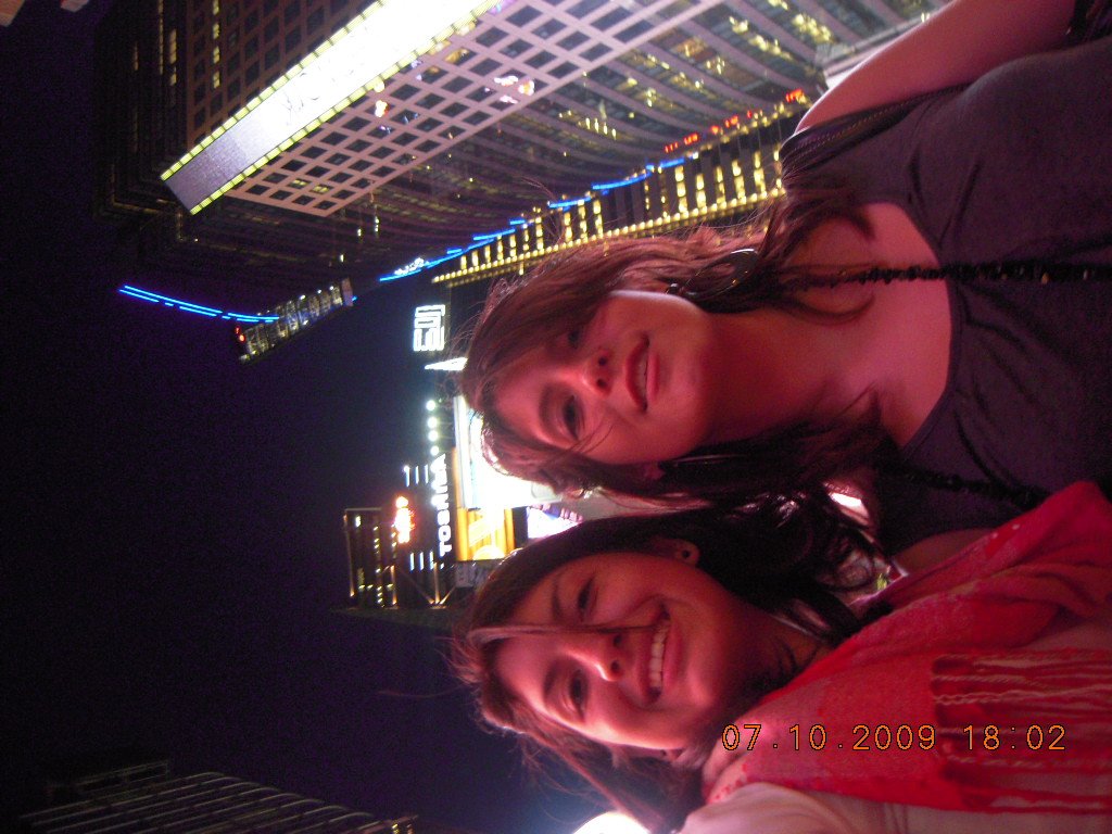 Two women smiling and posing for a photo at night in a city with bright building lights and neon signs.
