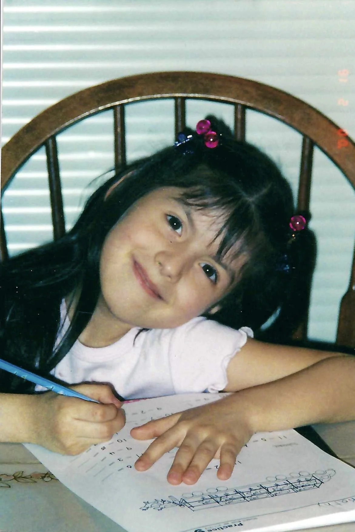 A young girl with dark hair and pink hair clips sitting at a table, smiling at the camera while doing homework with a pencil on a worksheet.