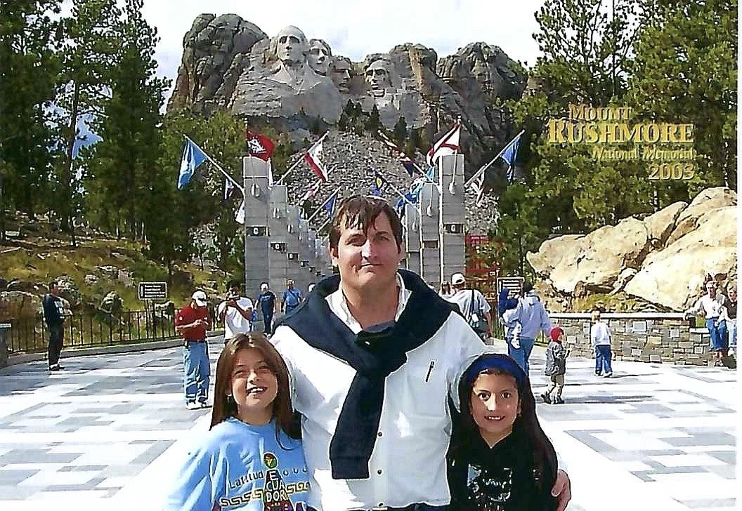 People standing in front of Mount Rushmore National Memorial with American flags, mountains, and trees in the background.