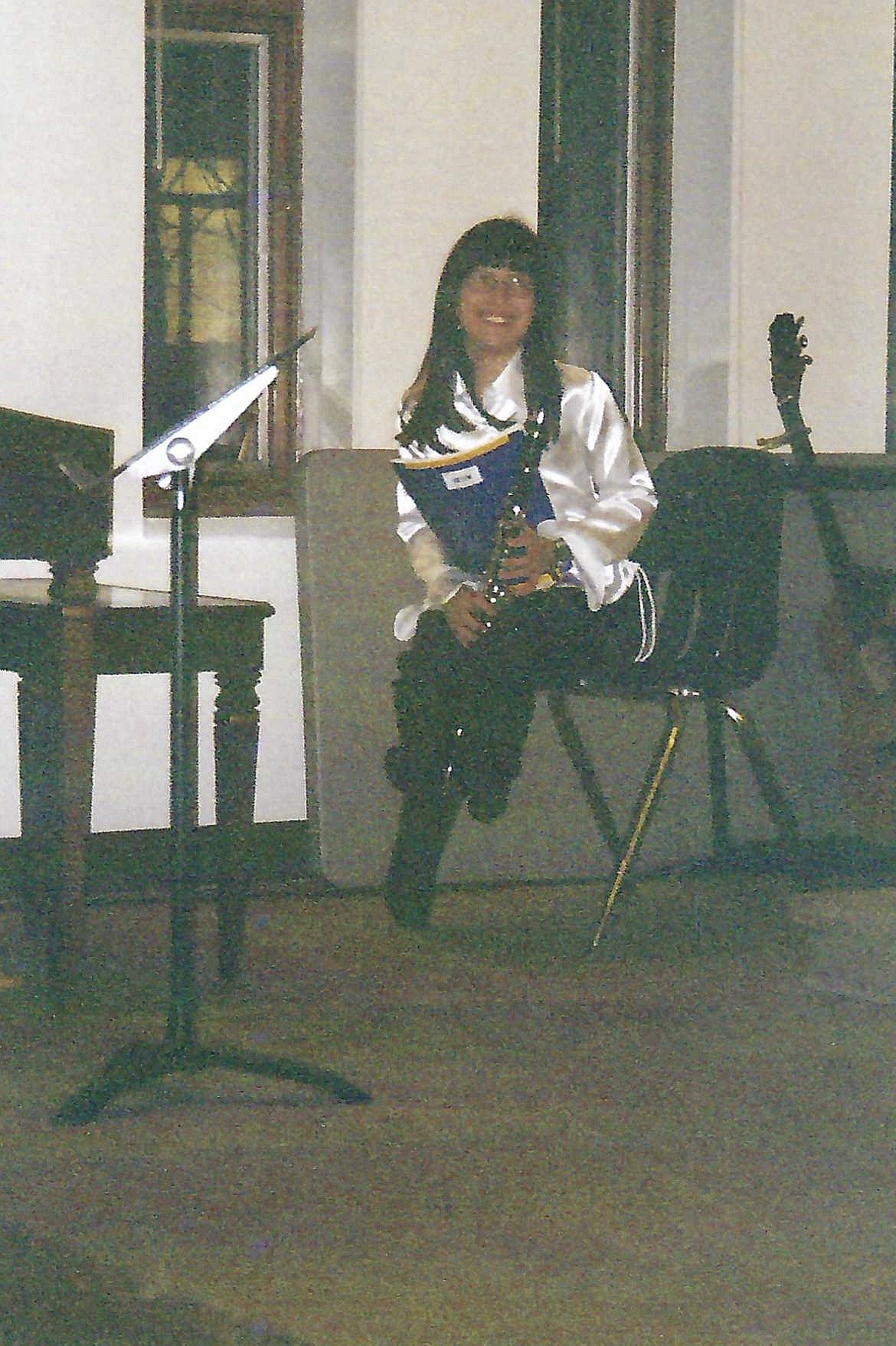 A woman with long black hair dressed in a white shirt, holding a folder, standing in a room with musical instruments, including a guitar and a microphone stand.